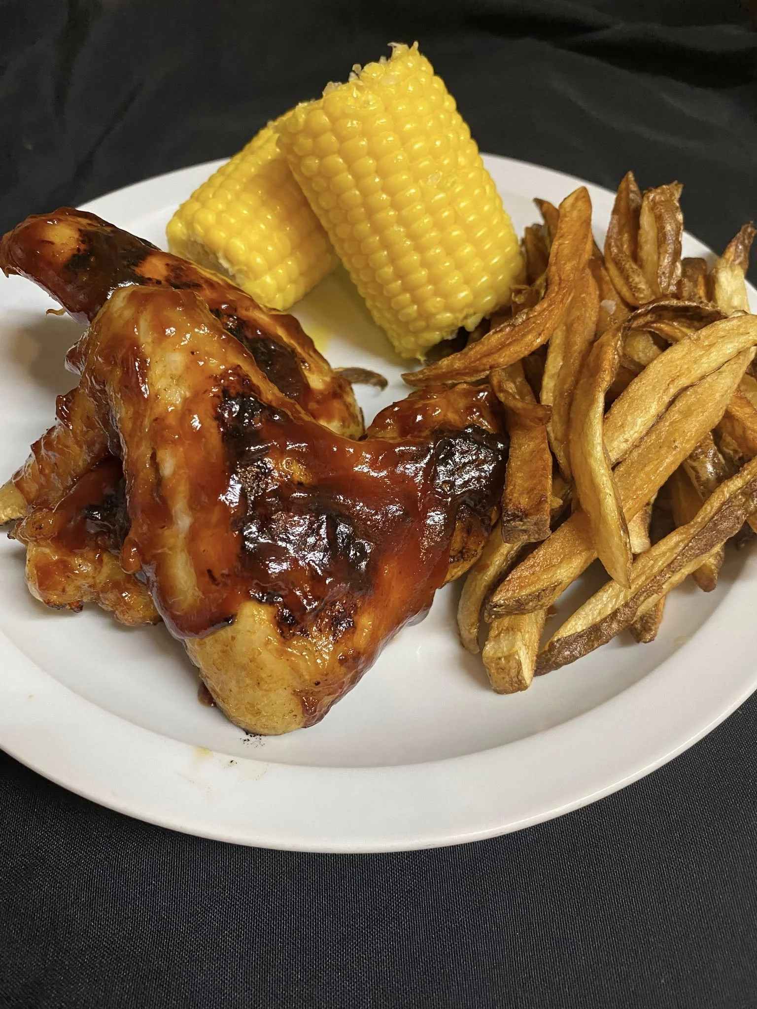 Plate of food with grilled corn, French fries, and barbecue chicken leg with sauce.