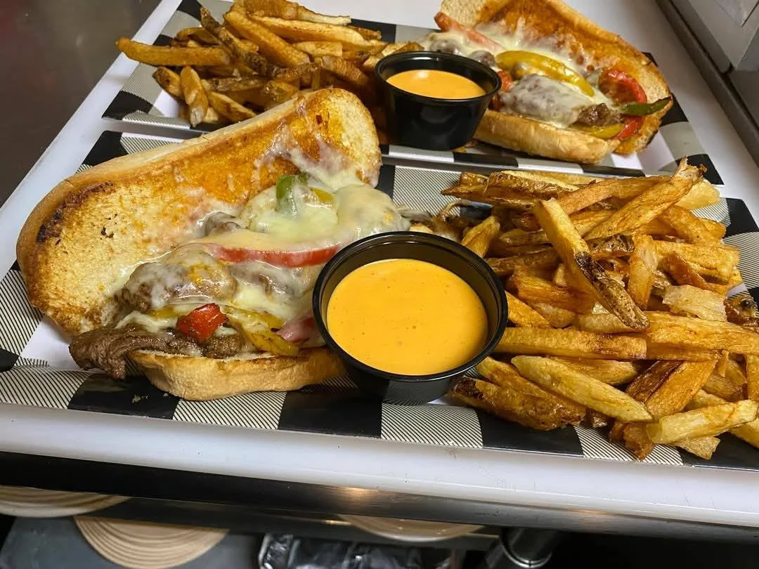 Two cheesesteak sandwiches with French fries and dipping sauces on a black and white checkered tray.