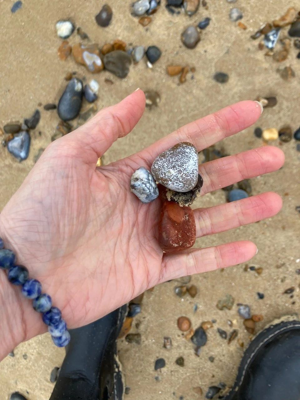 Hand holding four colorful rocks on sandy ground scattered with small stones, wearing a beaded bracelet and black boots.