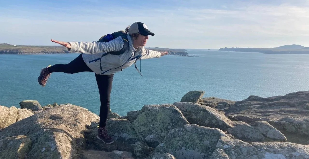 Woman balancing on rocks near the coast, with arms outstretched, wearing a cap, backpack, and casual clothes, overlooking the ocean and distant landforms.