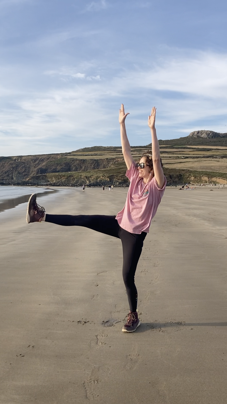 A woman in sunglasses smiling and balancing on one leg on the beach, with her arms raised and one leg extended out to the side.