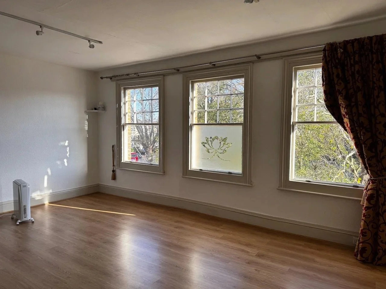 Empty room with three large windows, one partially covered by a patterned curtain, and a wooden floor.