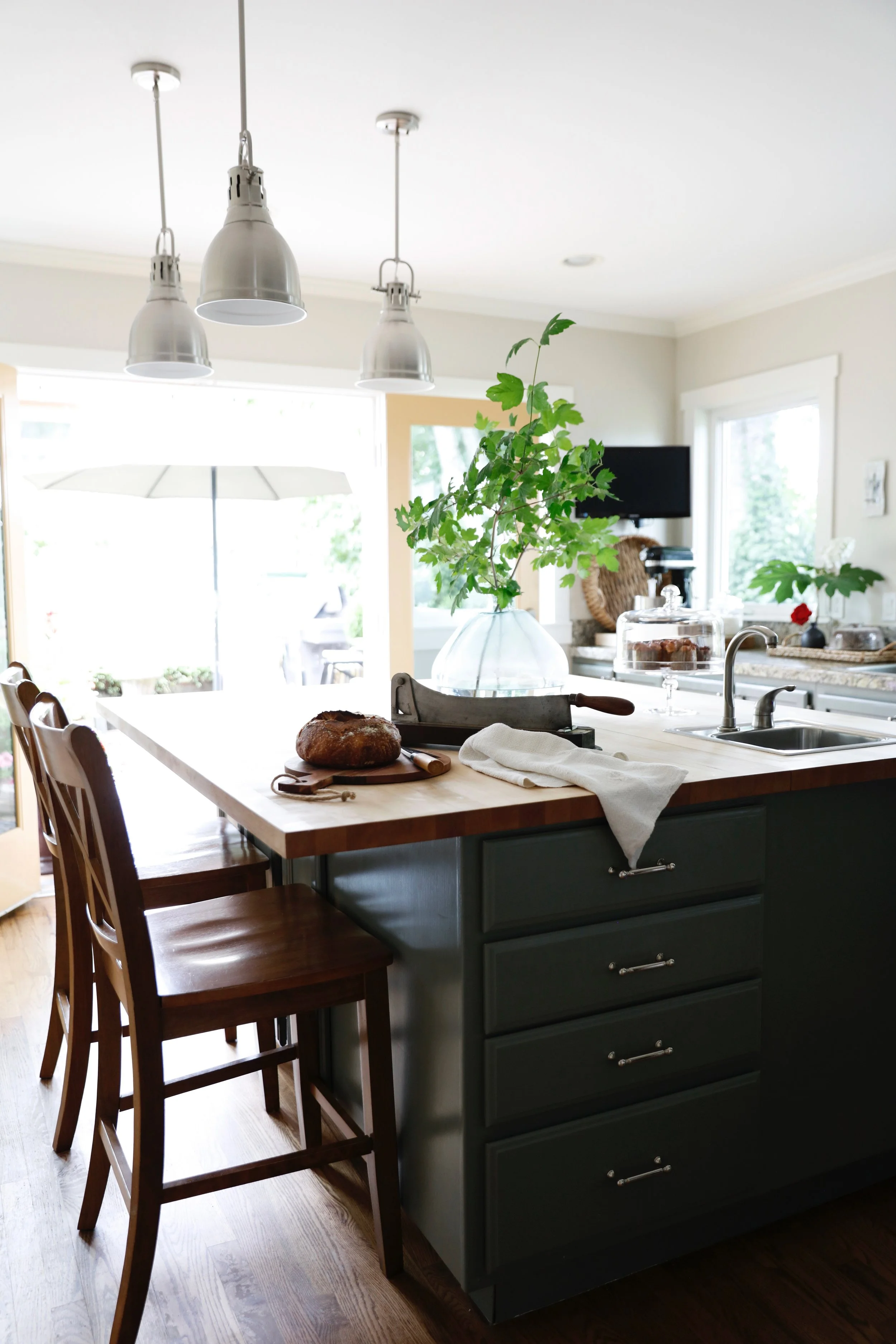 A kitchen island with a loaf of bread on a cutting board, a knife, a white towel, and a potted green plant. The island has dark drawers and a light wooden top. Three wooden chairs are on one side. In the background, a window, a television, and kitchen items are visible.