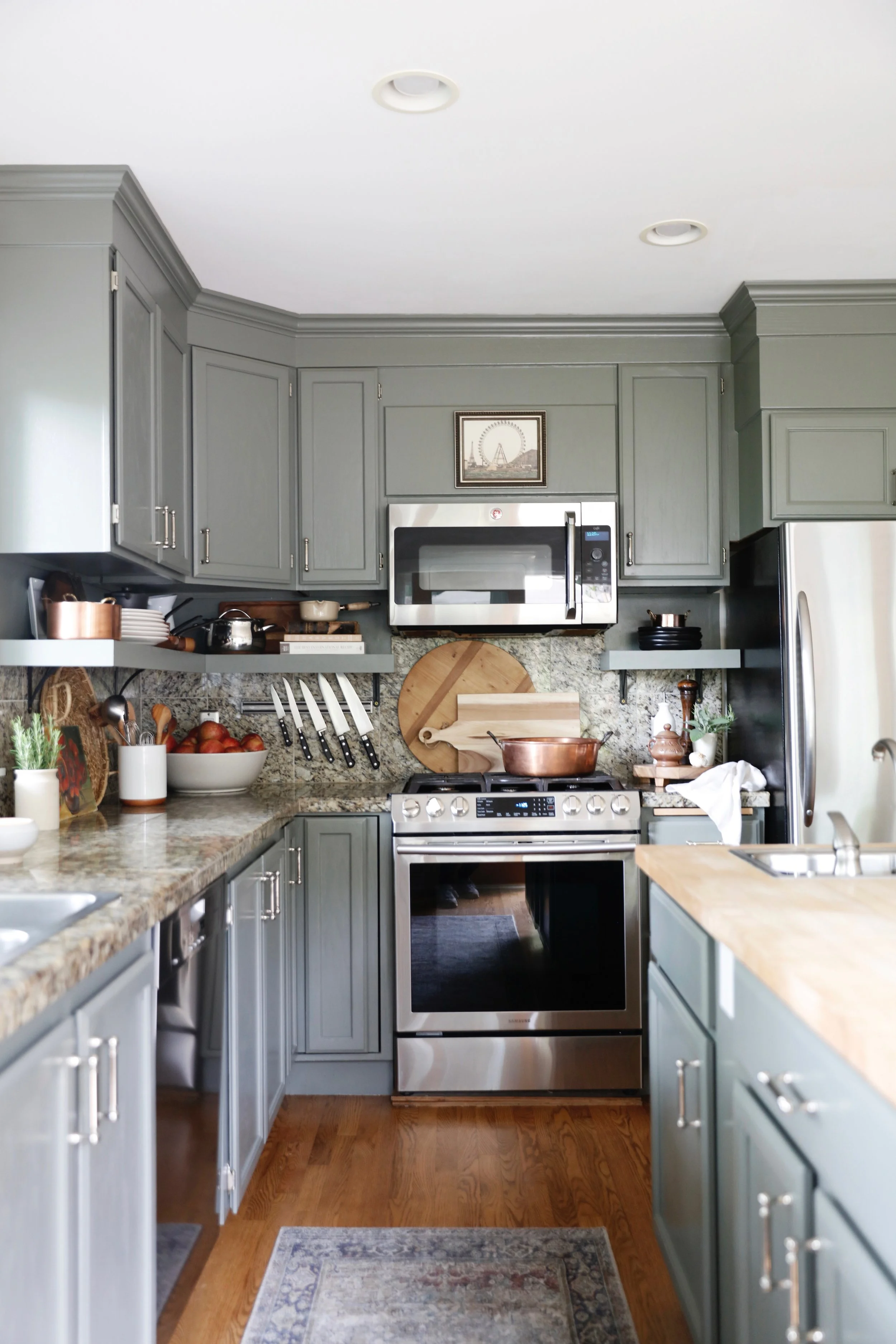 A kitchen with gray cabinets, a stainless steel stove and microwave, granite countertops, and wooden floor. Various kitchen utensils, bowls, and plants are visible.