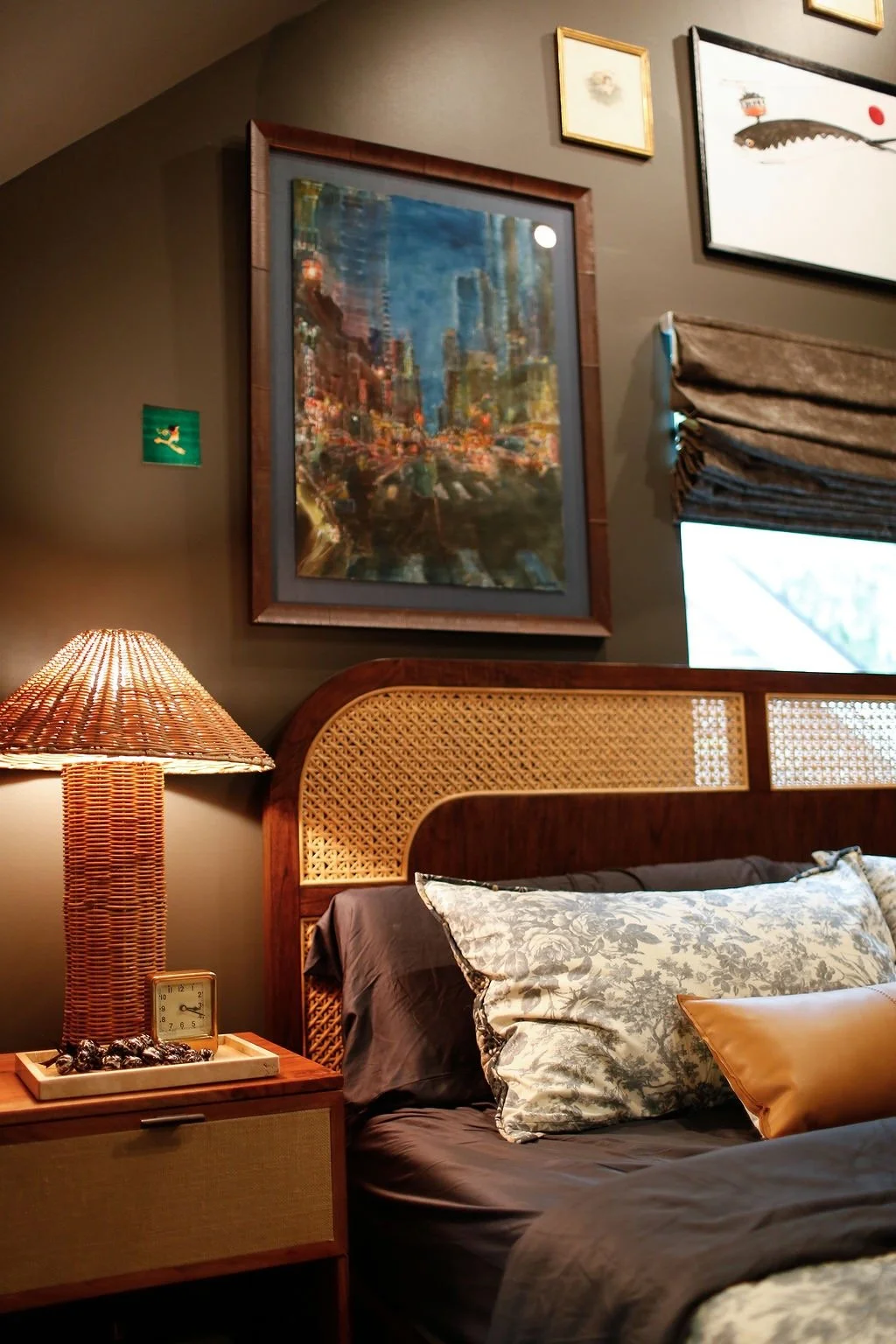 A cozy bedroom with a wooden bed frame, patterned pillows, a bedside lamp with a woven shade, a small clock, and various framed artwork on the wall.