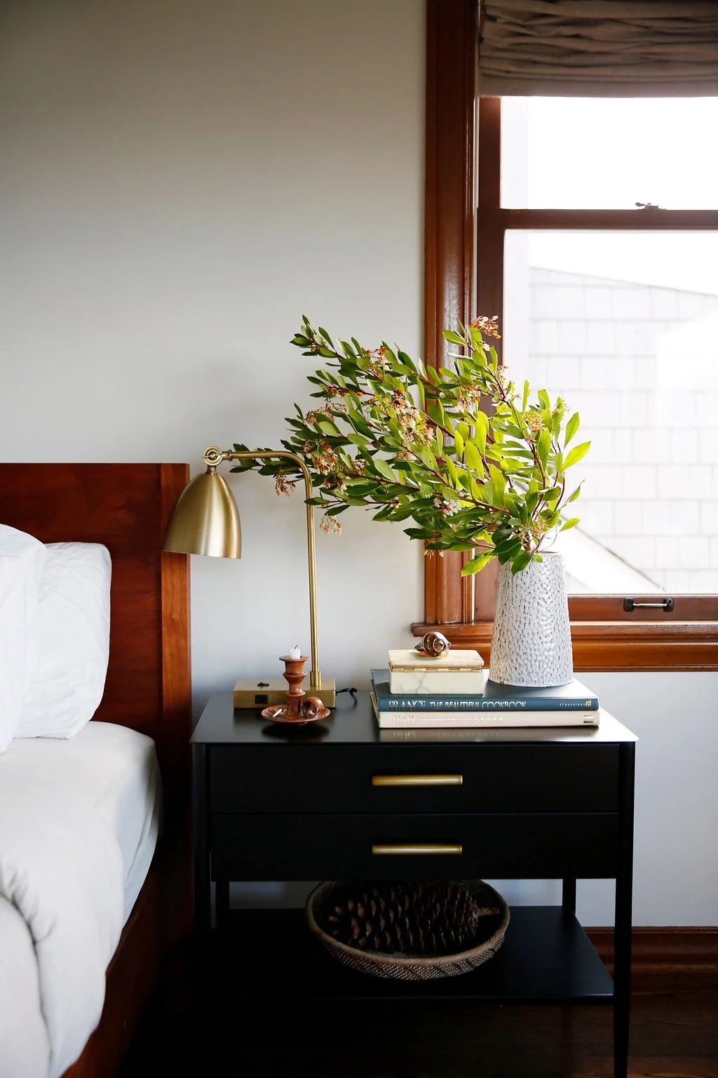 A bedroom side table with a gold desk lamp, a white vase with green foliage, books, and decorative objects, next to a window with wooden trim.
