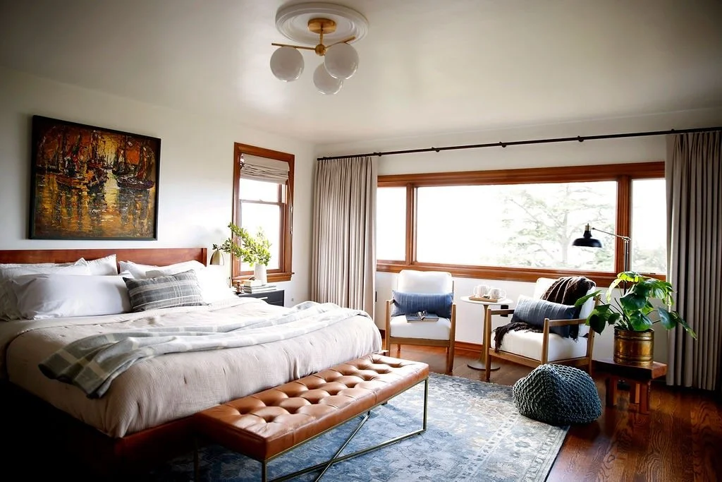 A cozy bedroom with a large bed, artwork above, and chairs near a wide window letting in natural light. Decor includes a potted plant, lamps, and a bench at the foot of the bed.