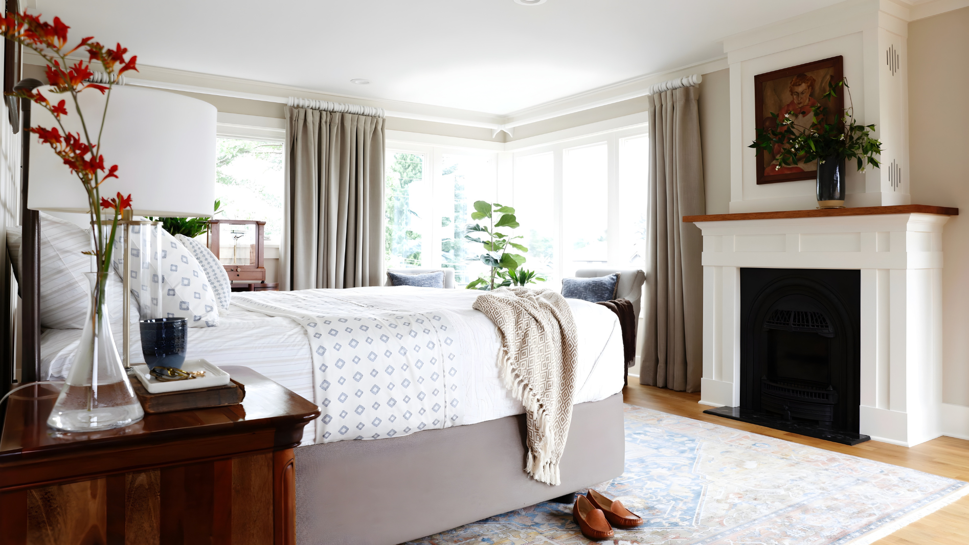 A cozy bedroom with a large bed featuring white and blue patterned bedding, located near a bay window with beige curtains. There is a wooden nightstand with a glass vase holding red flowers, a blue cup, and a tray. The room has a white fireplace with a wooden mantel decorated with a potted plant and a framed picture and a soft multicolored rug on the light wood floor. Natural light fills the room.