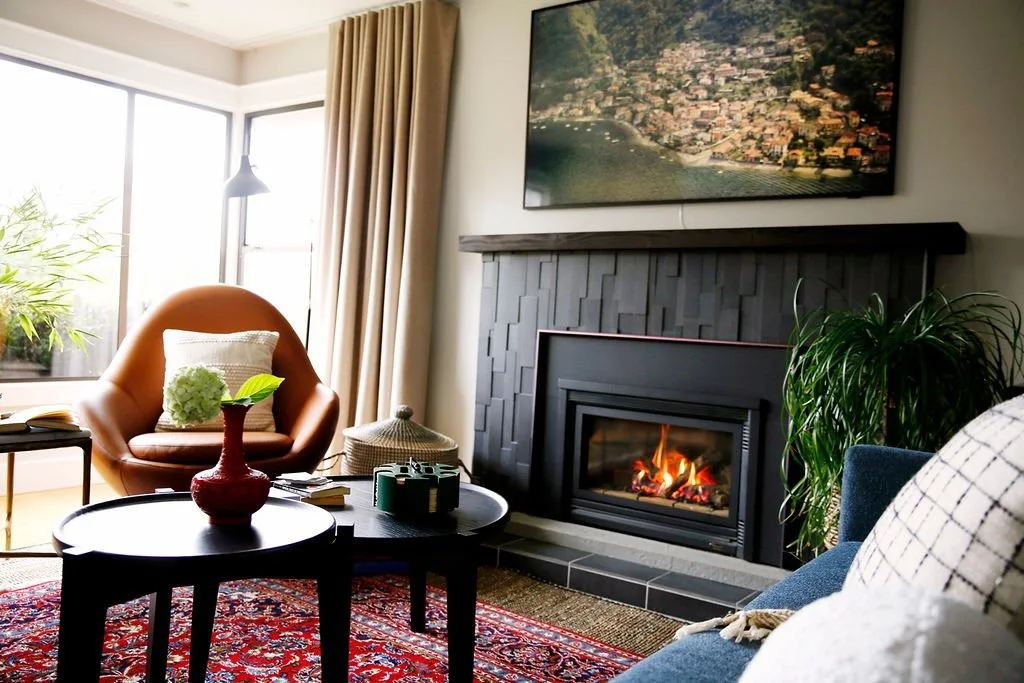 Living room with a fireplace, a large wall-mounted photo of a coastal town, a brown armchair with a white pillow, a black coffee table with decorative items, a blue sofa with patterned cushions, a potted plant, and large windows with beige curtains.