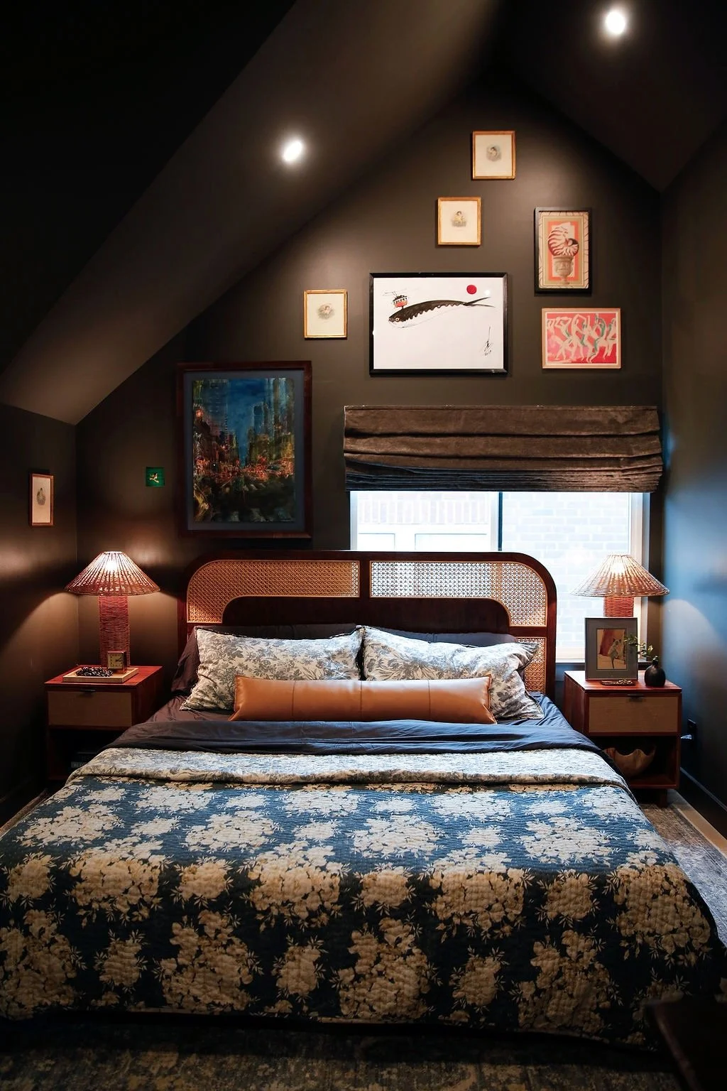 A cozy bedroom with a dark ceiling and walls, featuring a large bed with floral and leather pillows, two wooden nightstands with matching lamps, and various framed artwork on the wall above the bed, including paintings and illustrations.