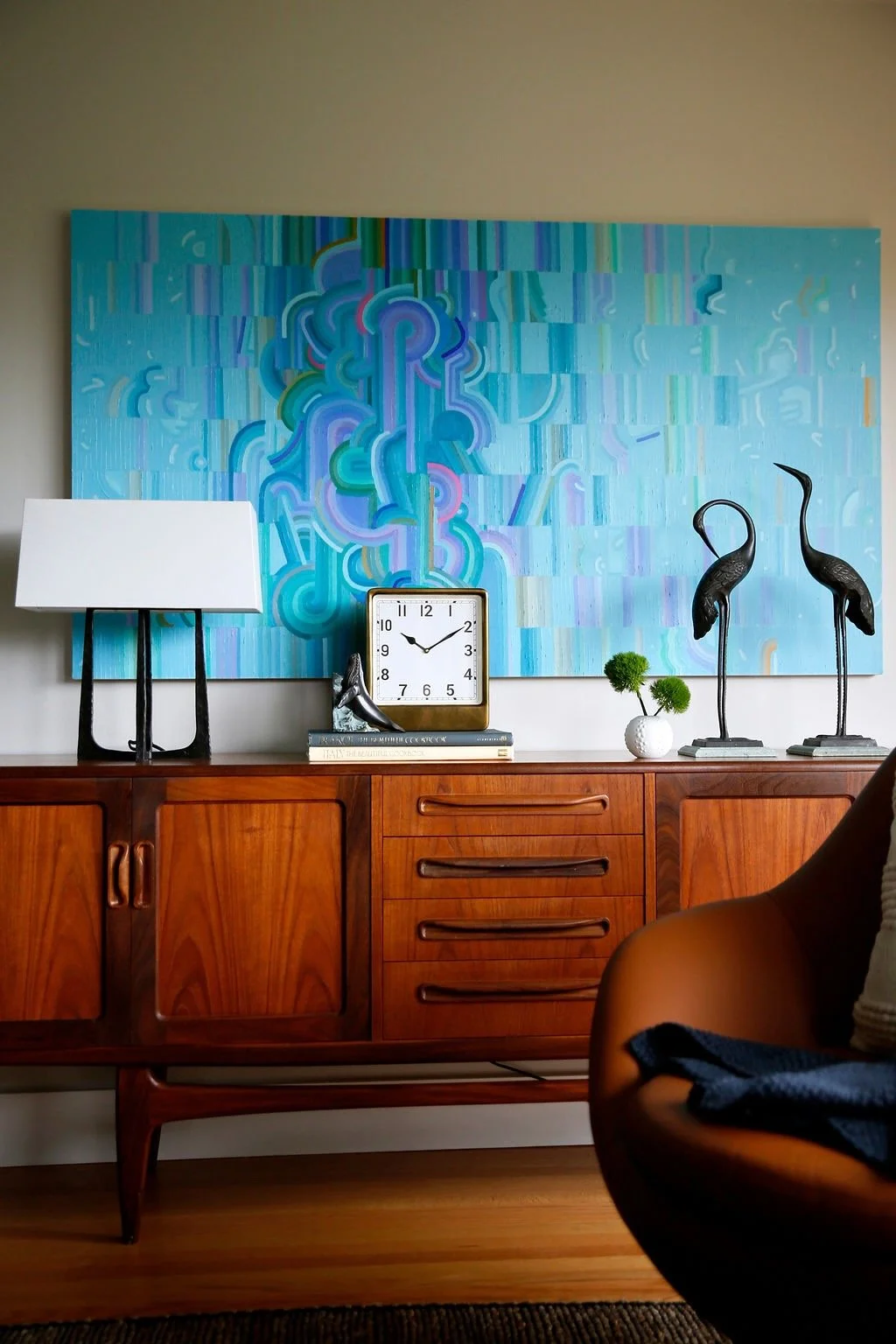 Mid-century modern wooden sideboard with decorative items including a black and white table lamp, a square clock, art books, two black crane sculptures, a small white vase with green plants, and an abstract blue and purple painting hanging on the wall behind.