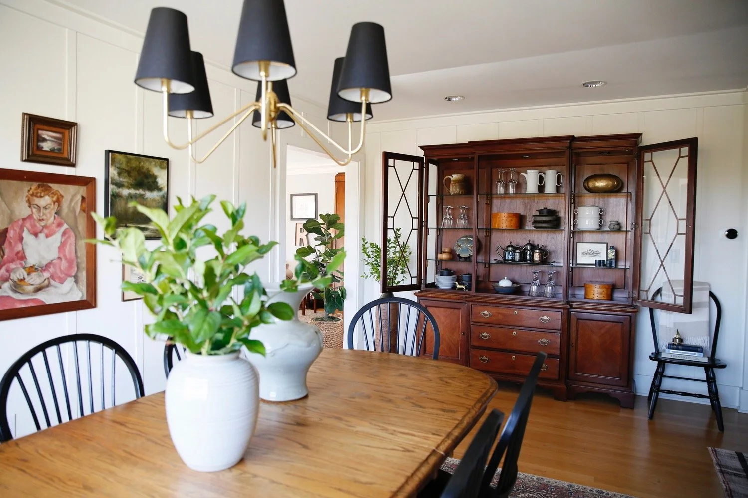 A dining room with a wooden table, black chairs, and a white vase with green plants. A wooden china cabinet with open doors displaying dishes and glasses. Artwork on the walls and a black chair with books in the corner.