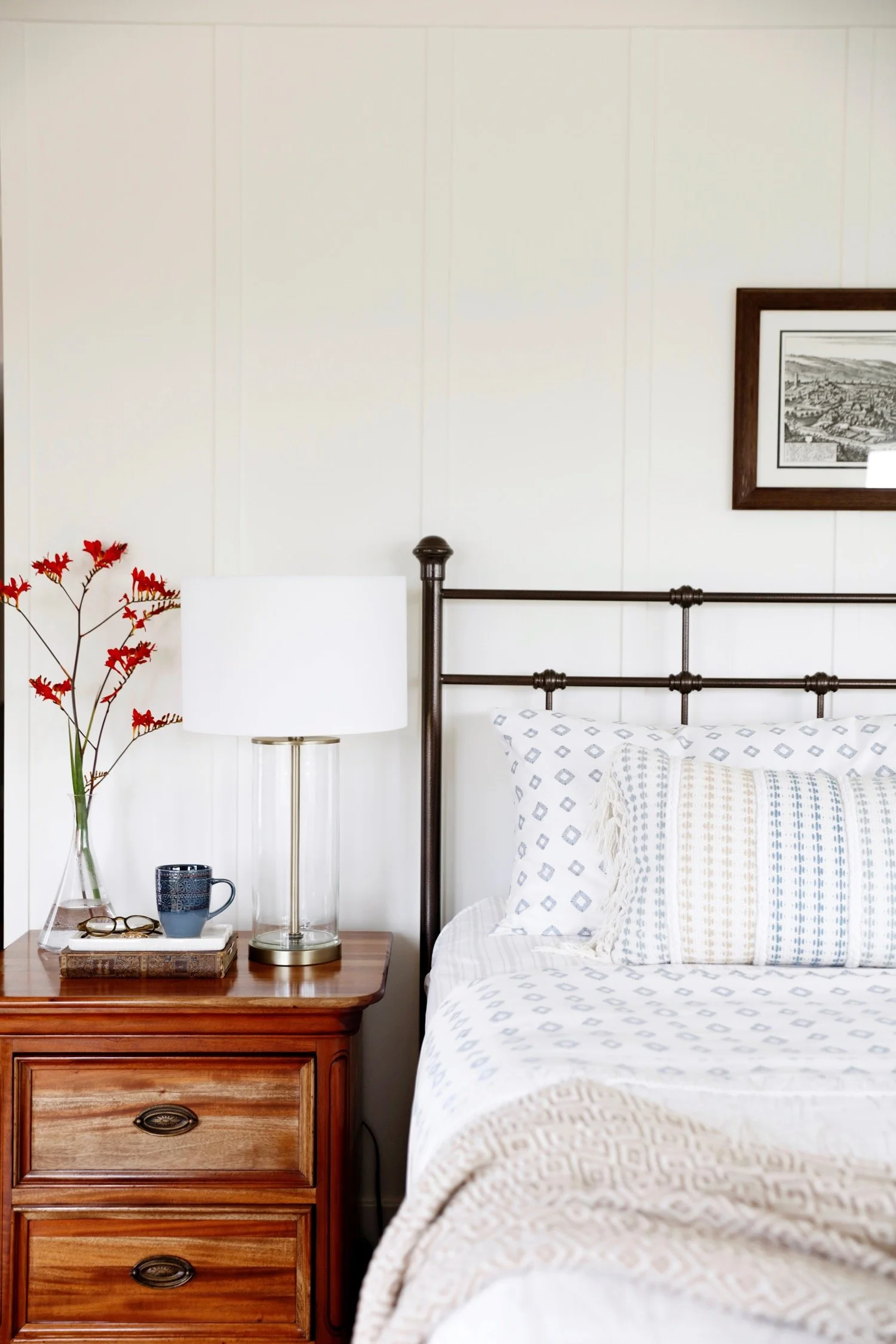 A bedroom with a wooden nightstand, a table lamp, a vase with red flowers, a cup, and glasses. Part of a bed with white and blue patterned bedding and a framed black-and-white picture on the wall.