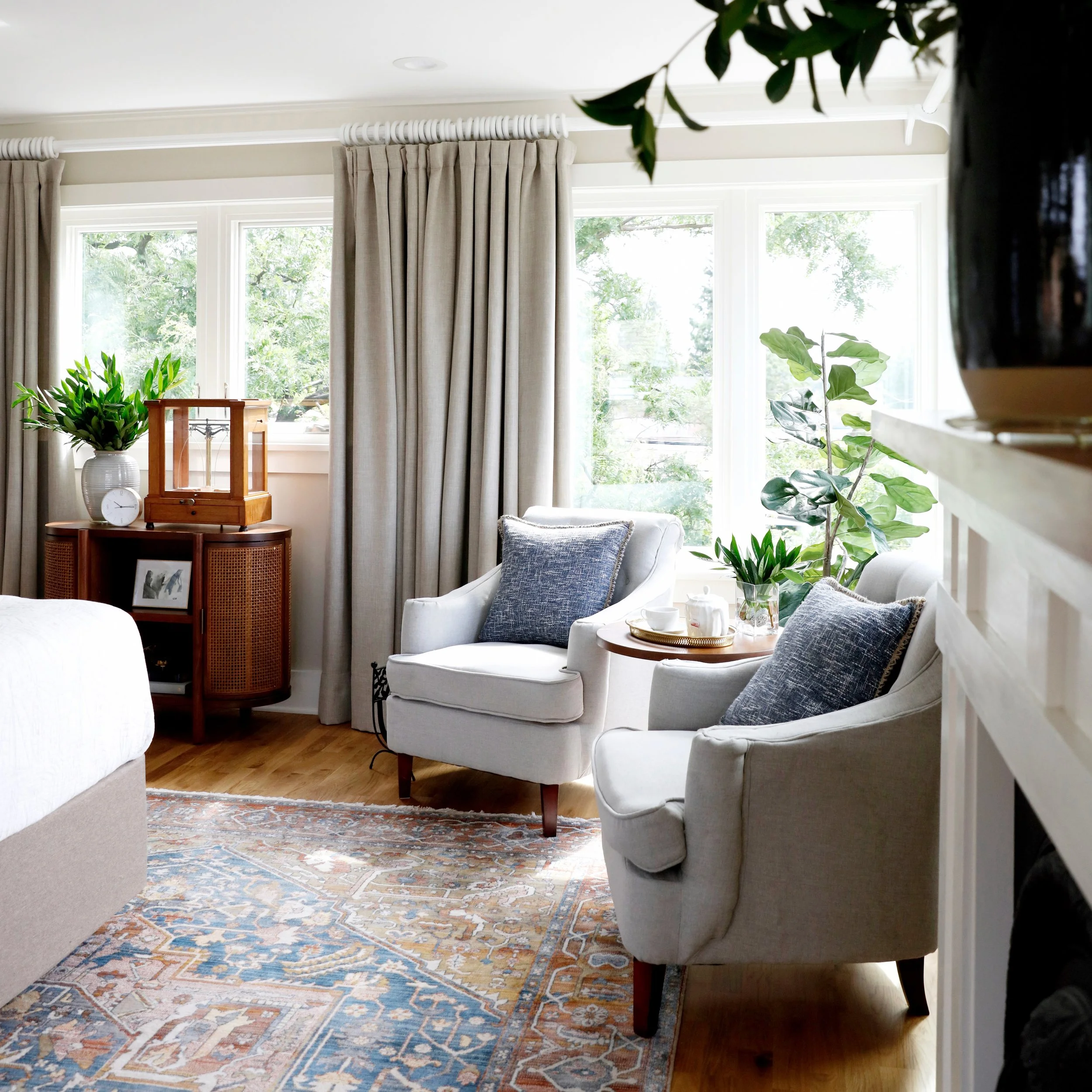 Living room with large windows, beige curtains, white armchair with blue pillows, a wooden side table with a tray, a potted plant, and a colorful rug.