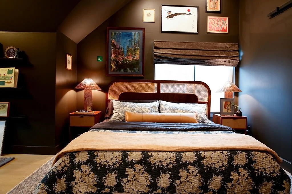 A bedroom with a king-sized bed, floral quilt, and decorative pillows, flanked by two wooden nightstands with lamps. The wall behind the bed has framed artwork and a window with a fabric shade.
