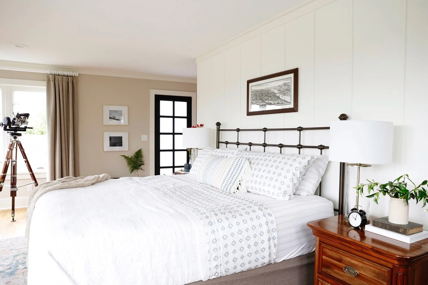 Bedroom with a made bed, white bedding, and decorative pillows. There are lamps on bedside tables, a framed picture on the wall, a telescope near the window, and a door with a black grid design.