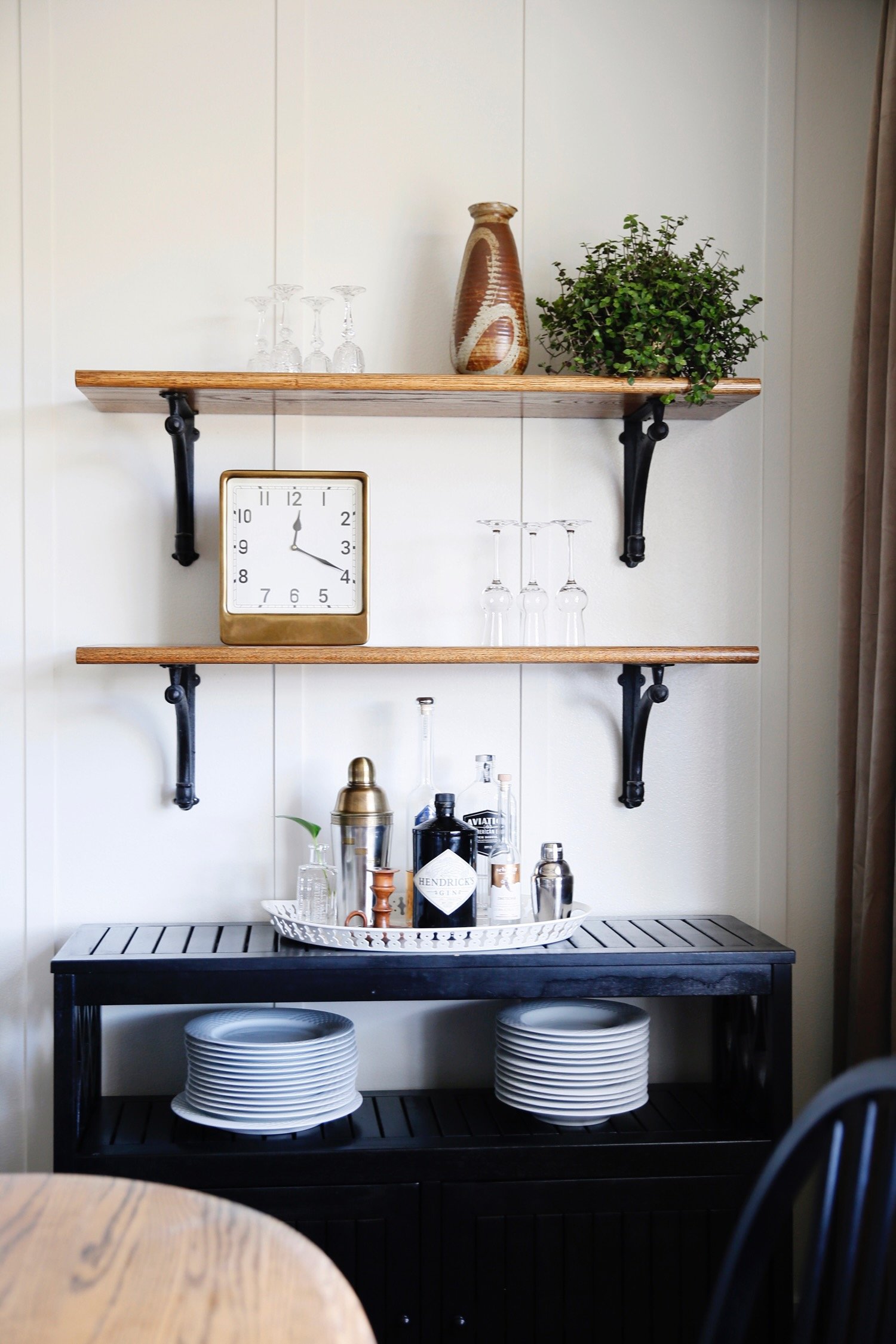 A black wooden sideboard with white plates, a tray with bottles and bar tools, two wooden shelves with glasses, a clock, a vase, and a potted plant on a white wall.