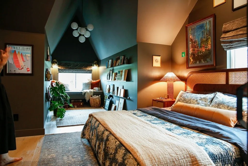 A cozy bedroom with a large bed, colorful pillows, a nightstand with a lamp, framed artwork on the wall, and a window with a roman shade. In the background, an alcove with a window seat, bookshelves, a large plant, a floor lamp, and framed pictures.