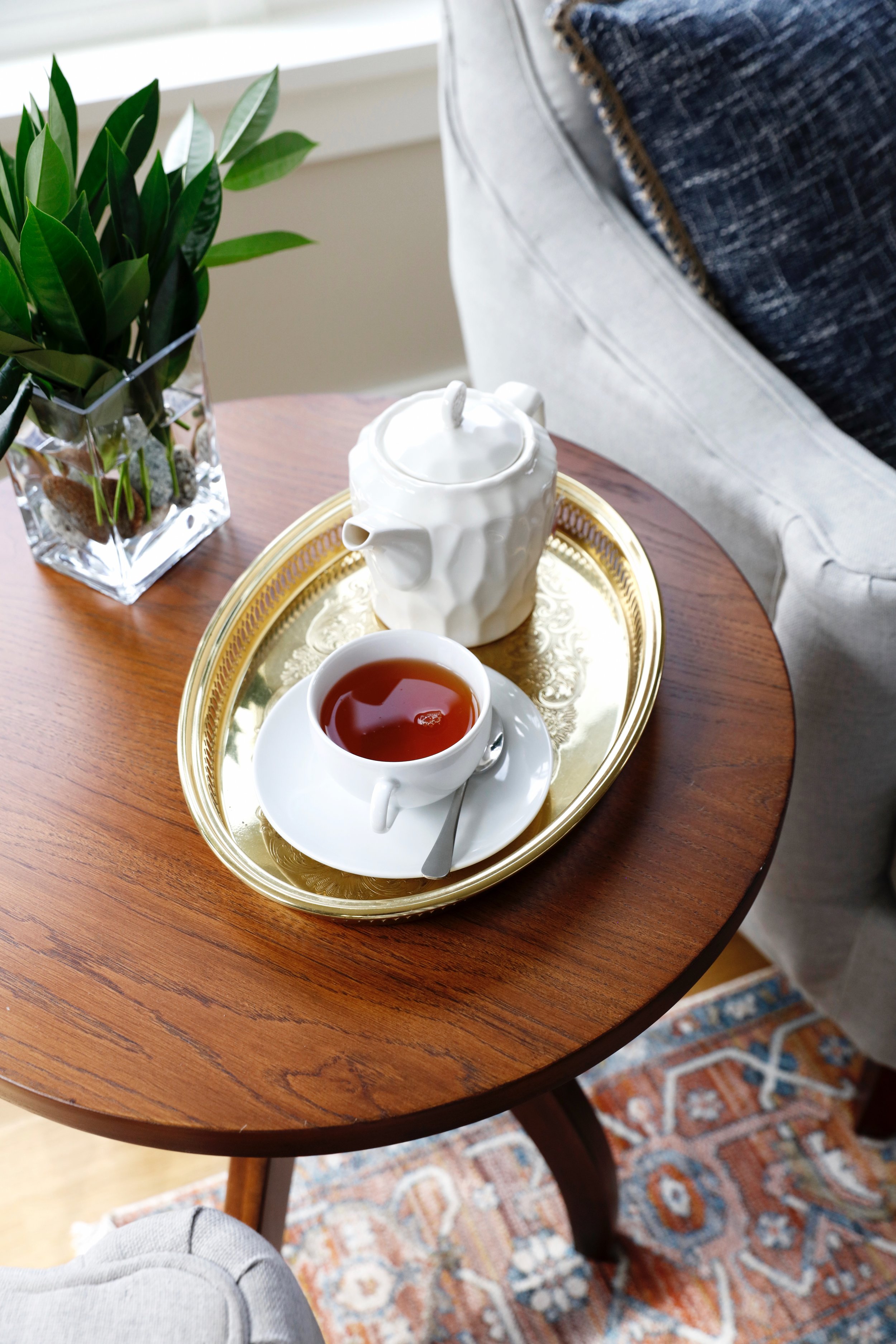 A living room scene featuring a wooden table with a gold tray holding a white teapot and a cup of tea. Next to the teapot, on the table, is a potted green plant in a clear glass vase. Part of a white upholstered armchair and a patterned rug are also visible.