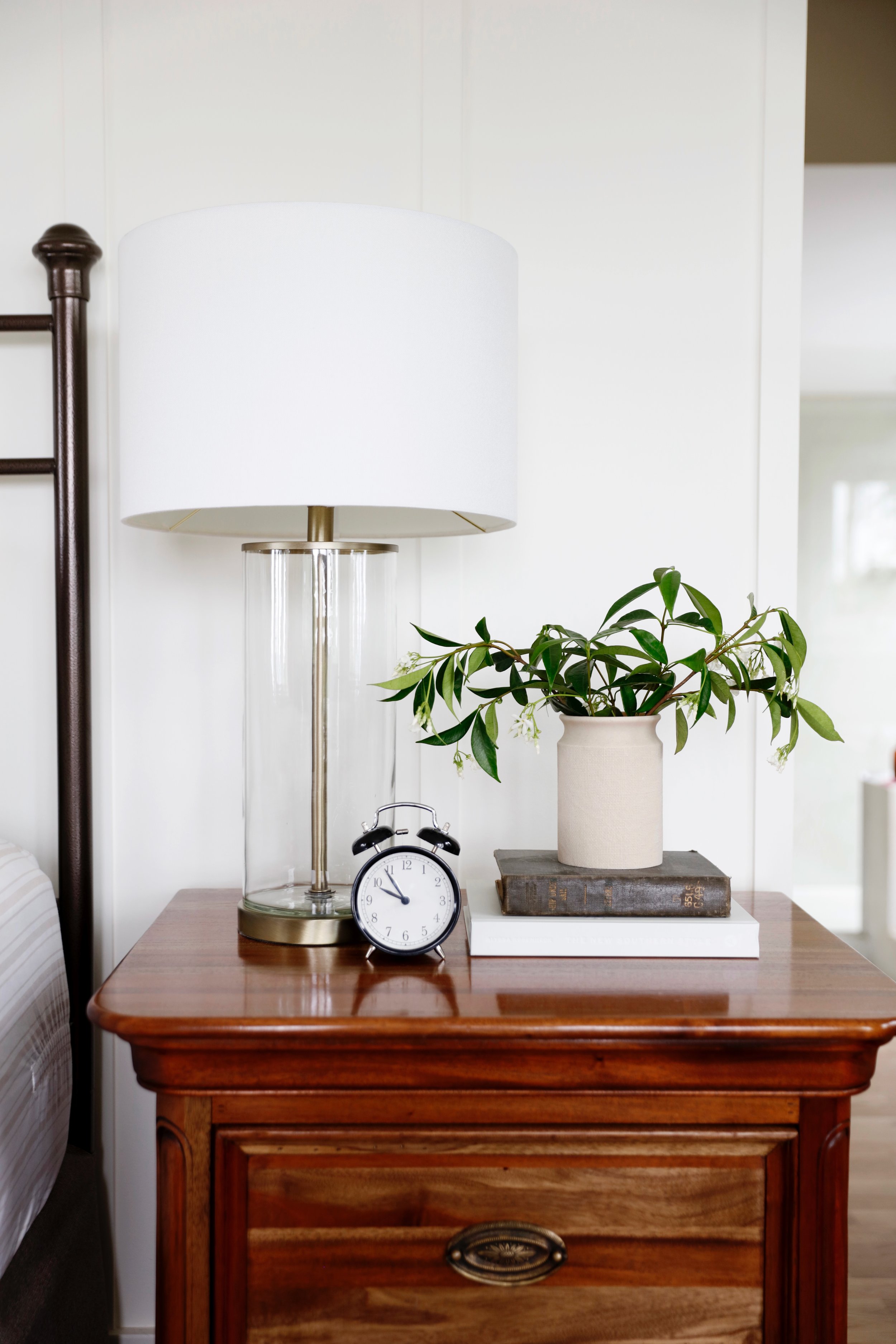 A wooden bedside table with a white lamp, a clock, a potted plant, and a book on top, against a white wall.
