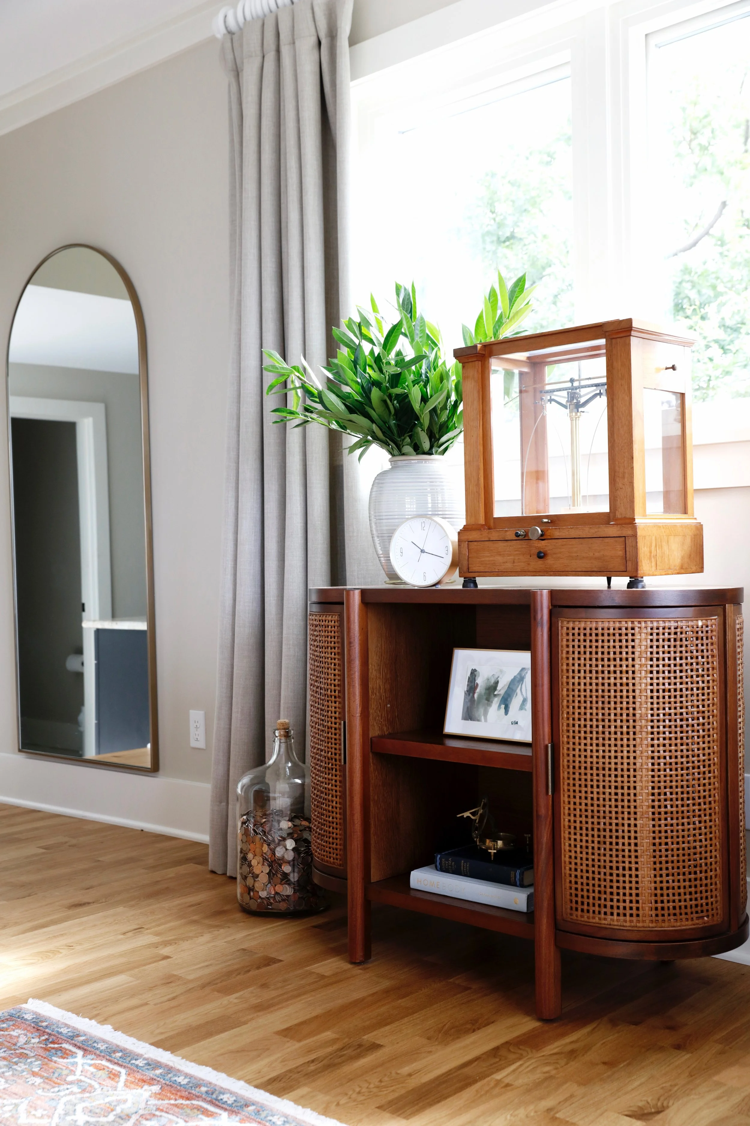A wooden and rattan cabinet with decorative items on top, including a glass case, a white clock, and a potted plant. The cabinet is next to a large window with beige curtains, and a mirror is on the wall nearby. A large jar filled with coins is on the floor beside the cabinet.