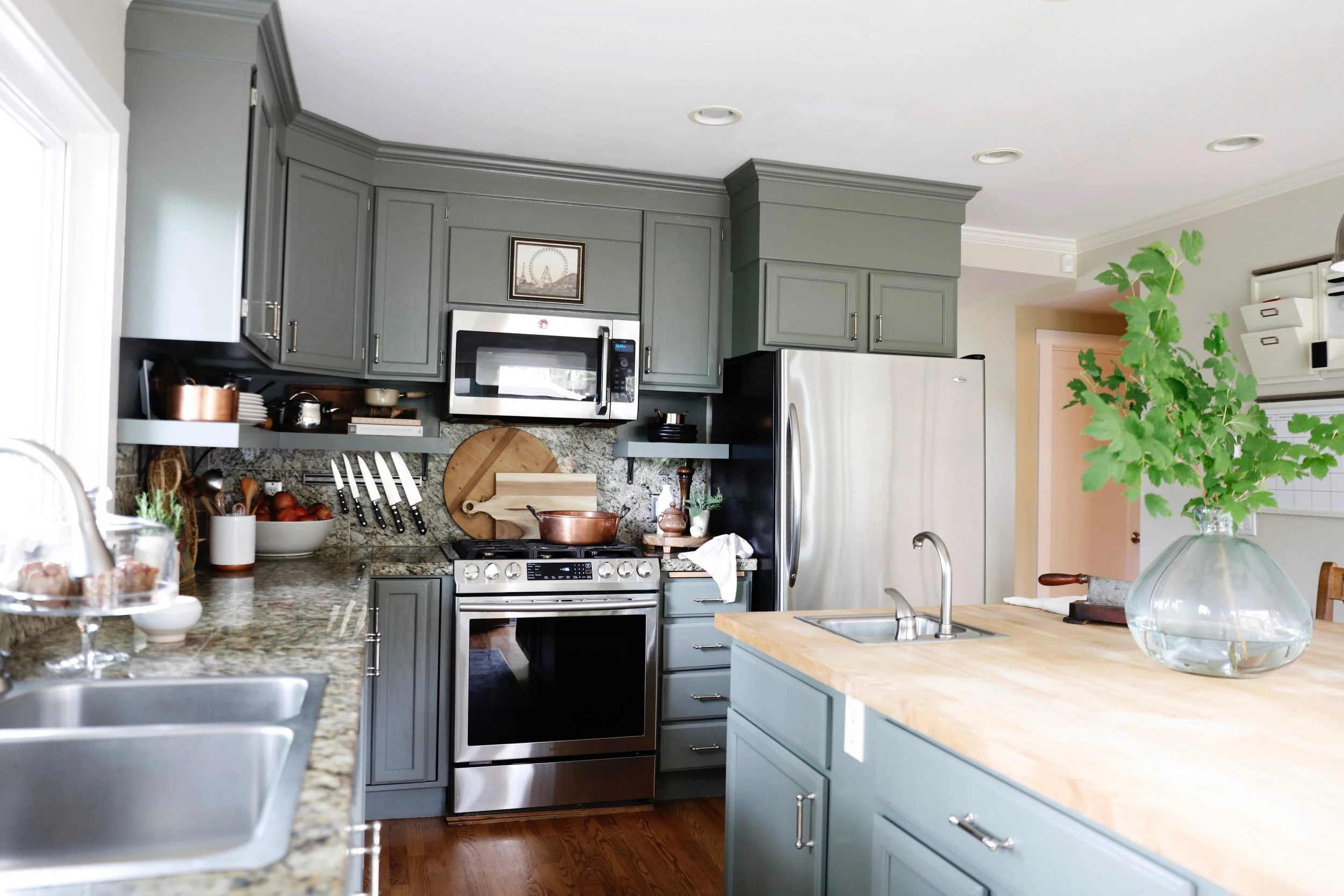 A modern kitchen with gray cabinets, a stainless steel microwave, a gas stove, granite countertops, a cutting board, a large green houseplant, and a small glass cake stand on the counter.
