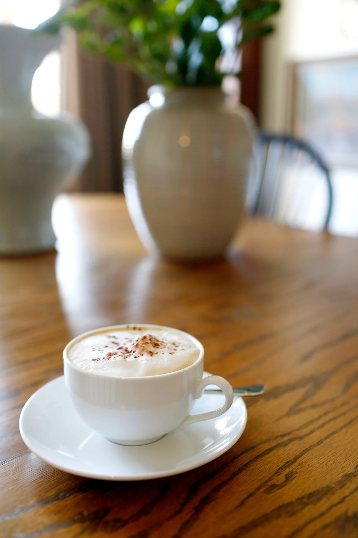 A close-up of a cup of cappuccino with foam, sprinkled with cocoa powder, on a white saucer with a spoon, placed on a wooden table.