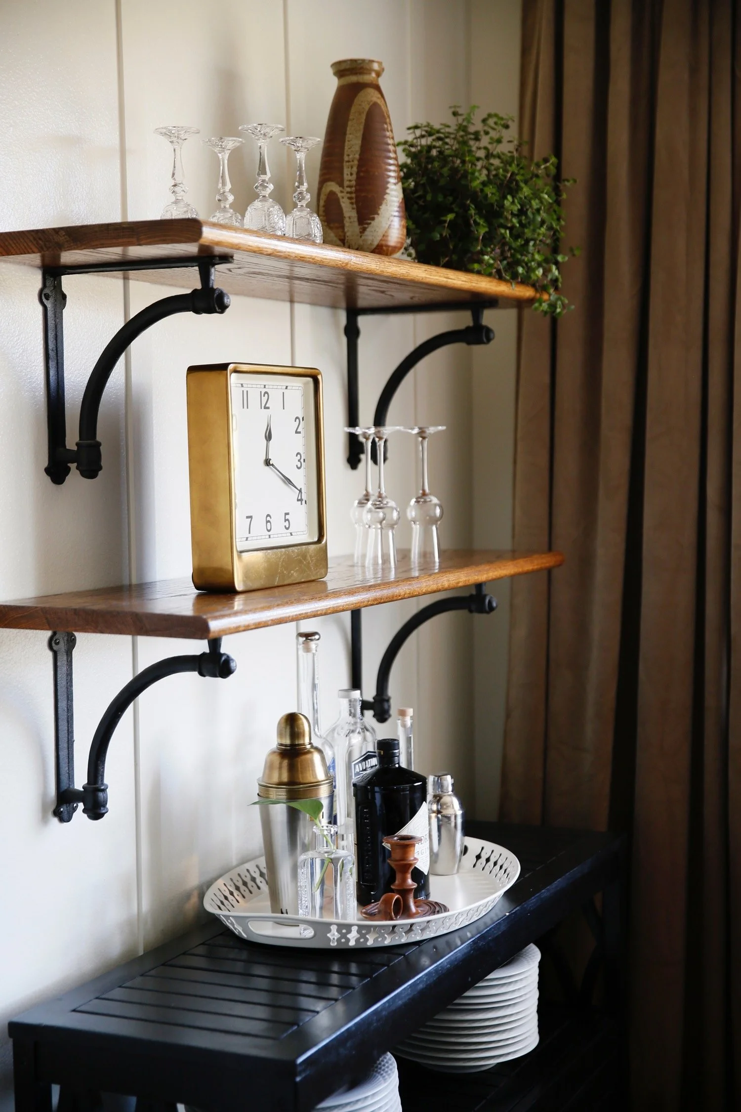 Decorative shelves with glassware, a gold clock, and a potted plant, set against a wall with curtains.