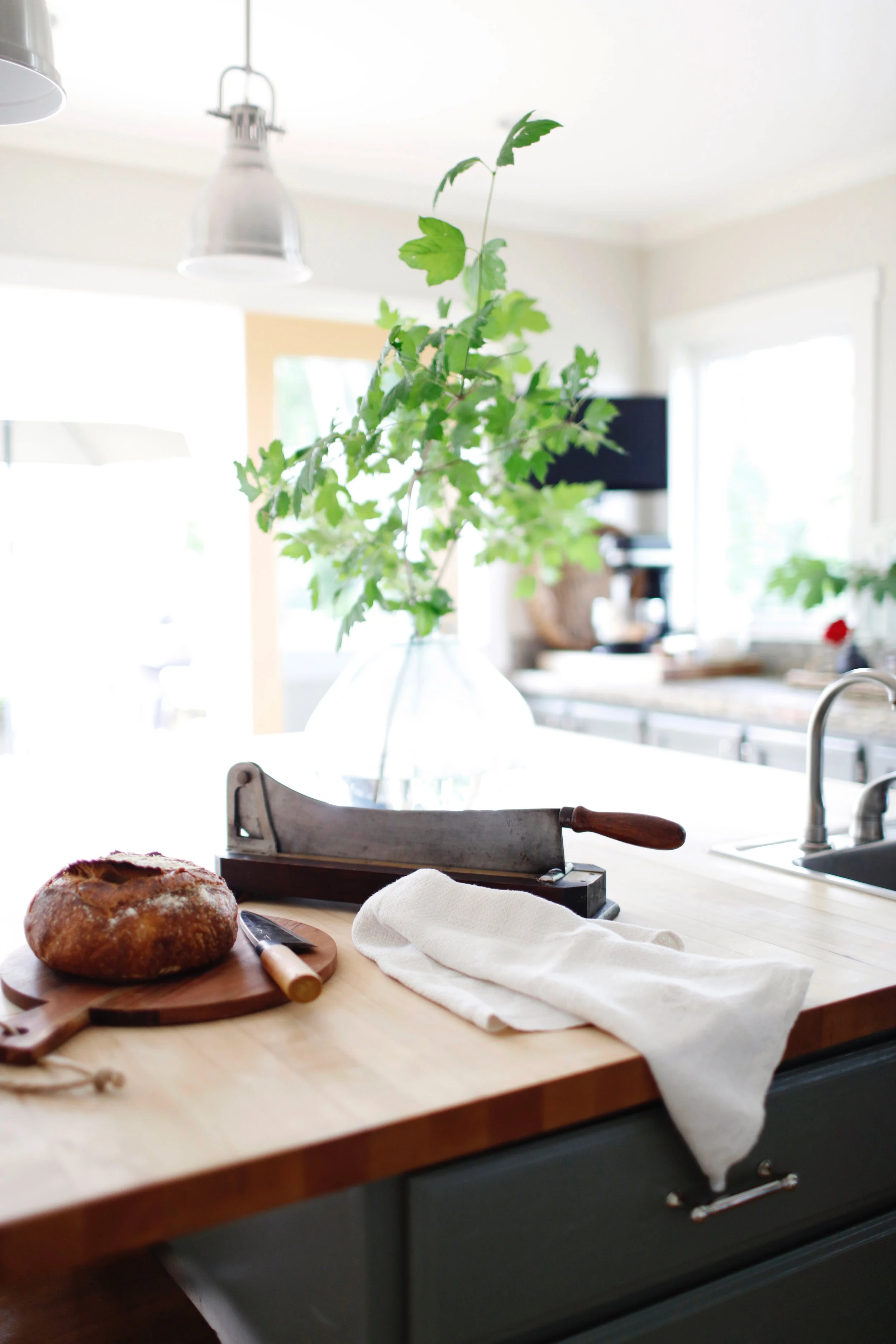 Kitchen with a loaf of bread on a wooden cutting board, a bread knife, a meat cleaver, a white cloth, a vase with green leafy branches, and a sink, in a bright and airy space.