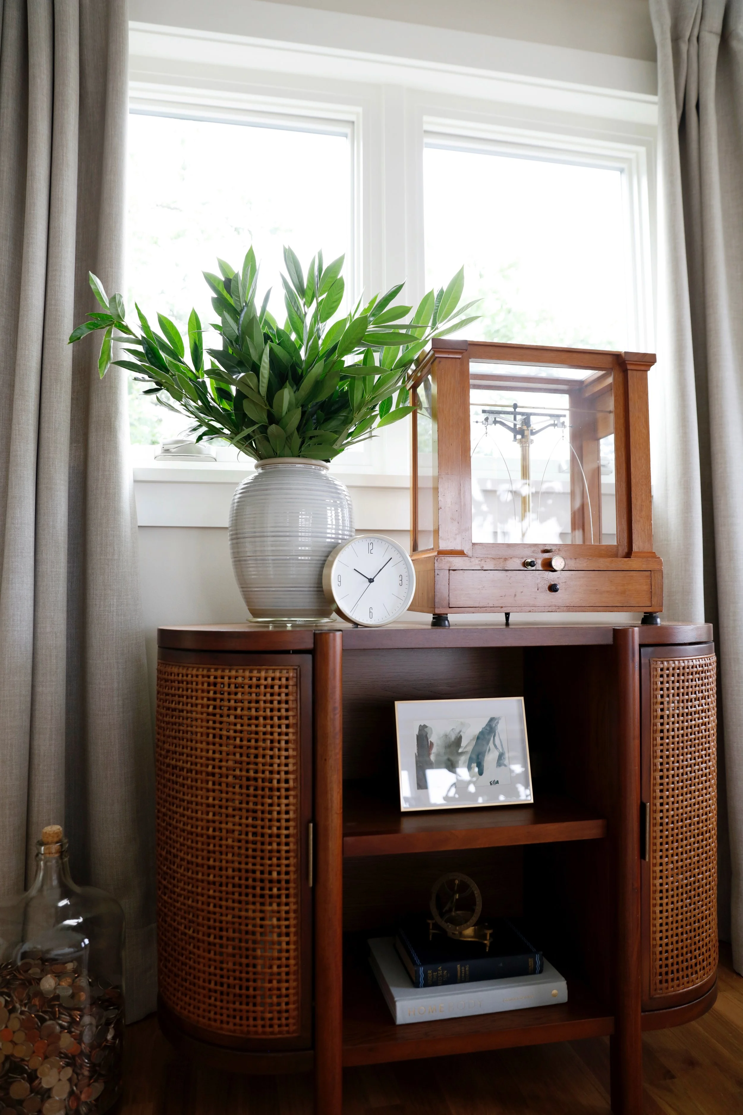 A wooden cabinet with rattan-wrapped sides, a glass clock, a framed abstract artwork, and a wooden supply box on top, with a large green plant in a white pot and a window with beige curtains behind it.