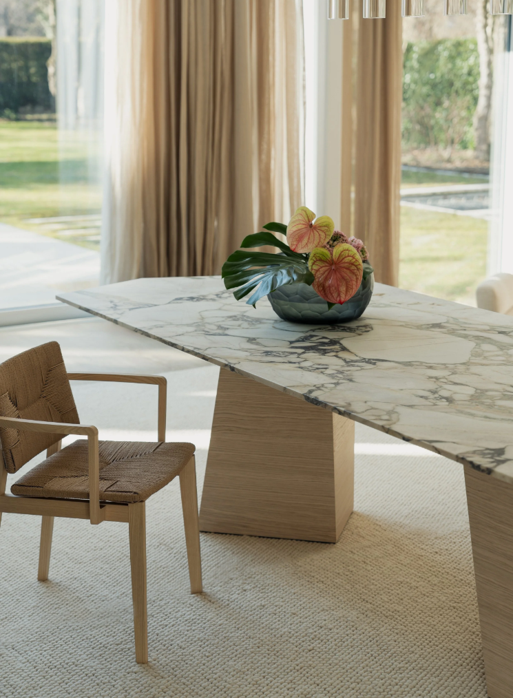 A dining room table with a white marble top and a wooden base, adorned with a floral arrangement in a dark vase, located near large windows with beige curtains, and a woven chair with wooden armrests positioned beside the table.