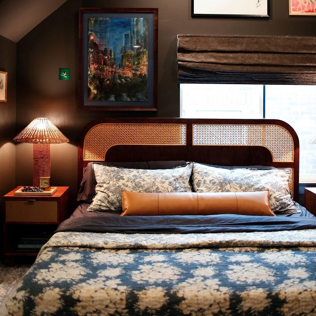 A cozy bedroom with a large bed, patterned bedding, decorative pillows, a wooden headboard with a woven design, a nightstand with a lamp, and framed artwork on a dark wall.