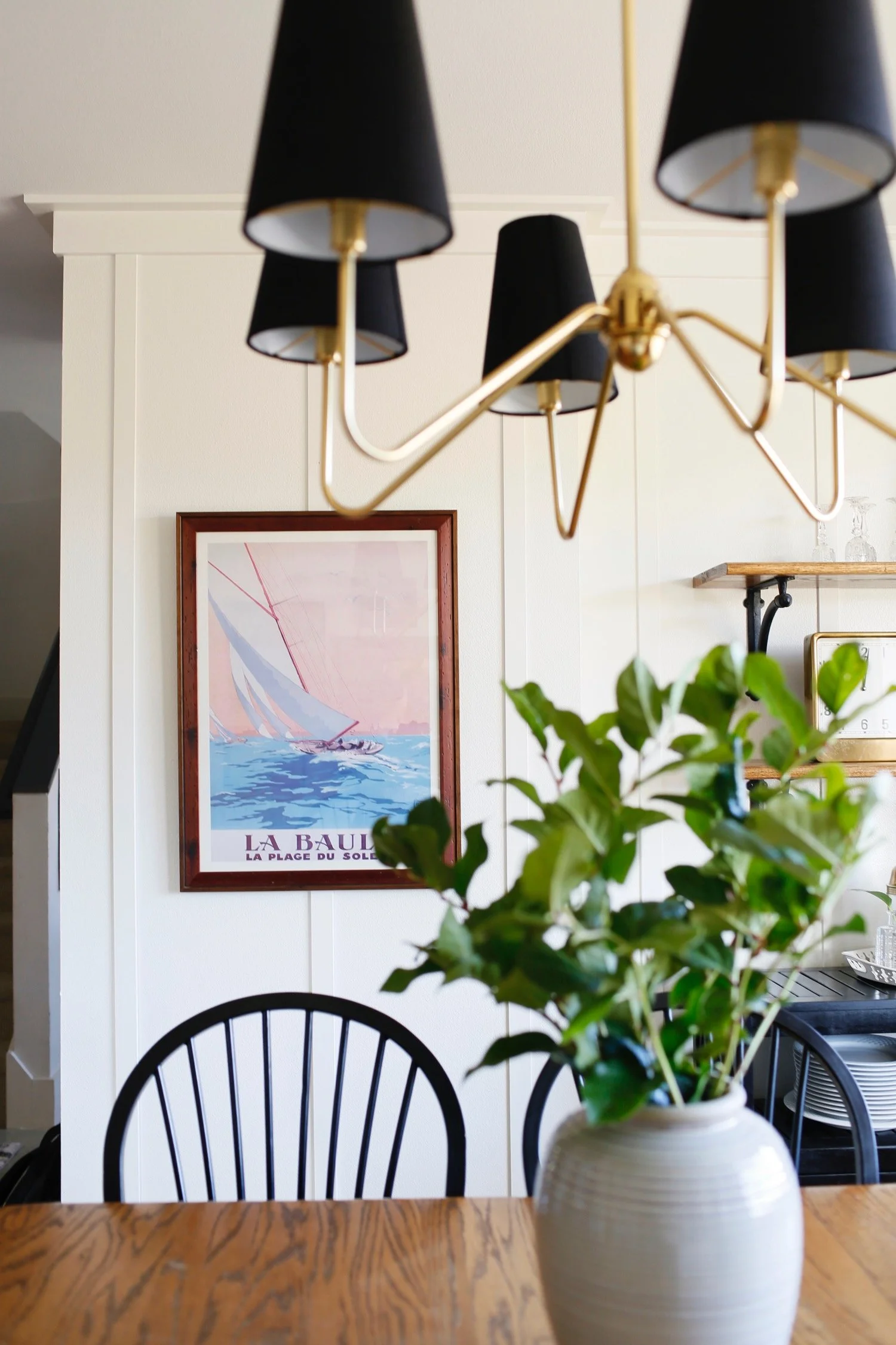 A dining area with a vase of green leafy plants on a wooden table, black chairs, and a framed picture of a sailboat on the wall.