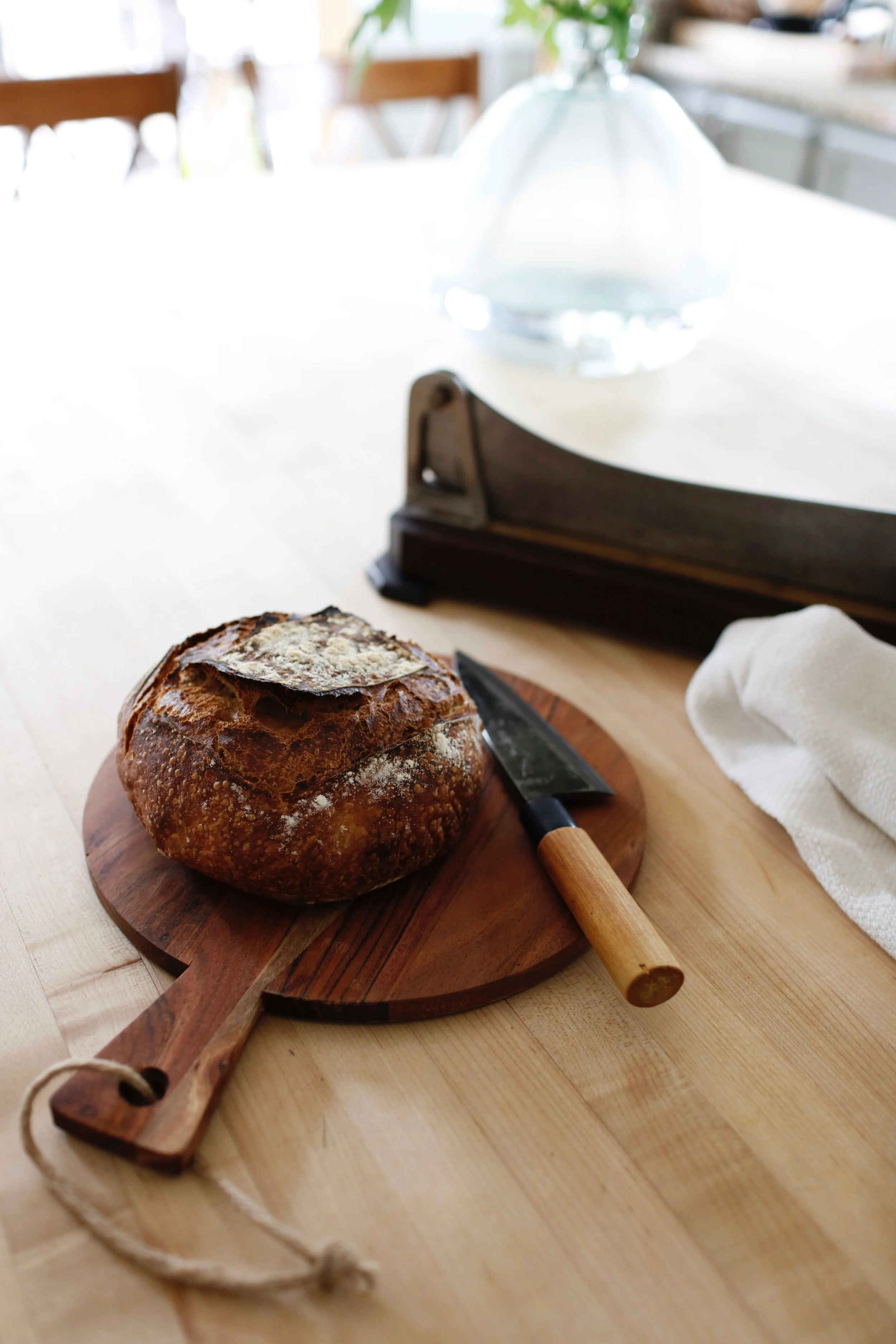 A loaf of rustic bread on a wooden cutting board next to a bread knife and a white cloth napkin on a light-colored wooden table.