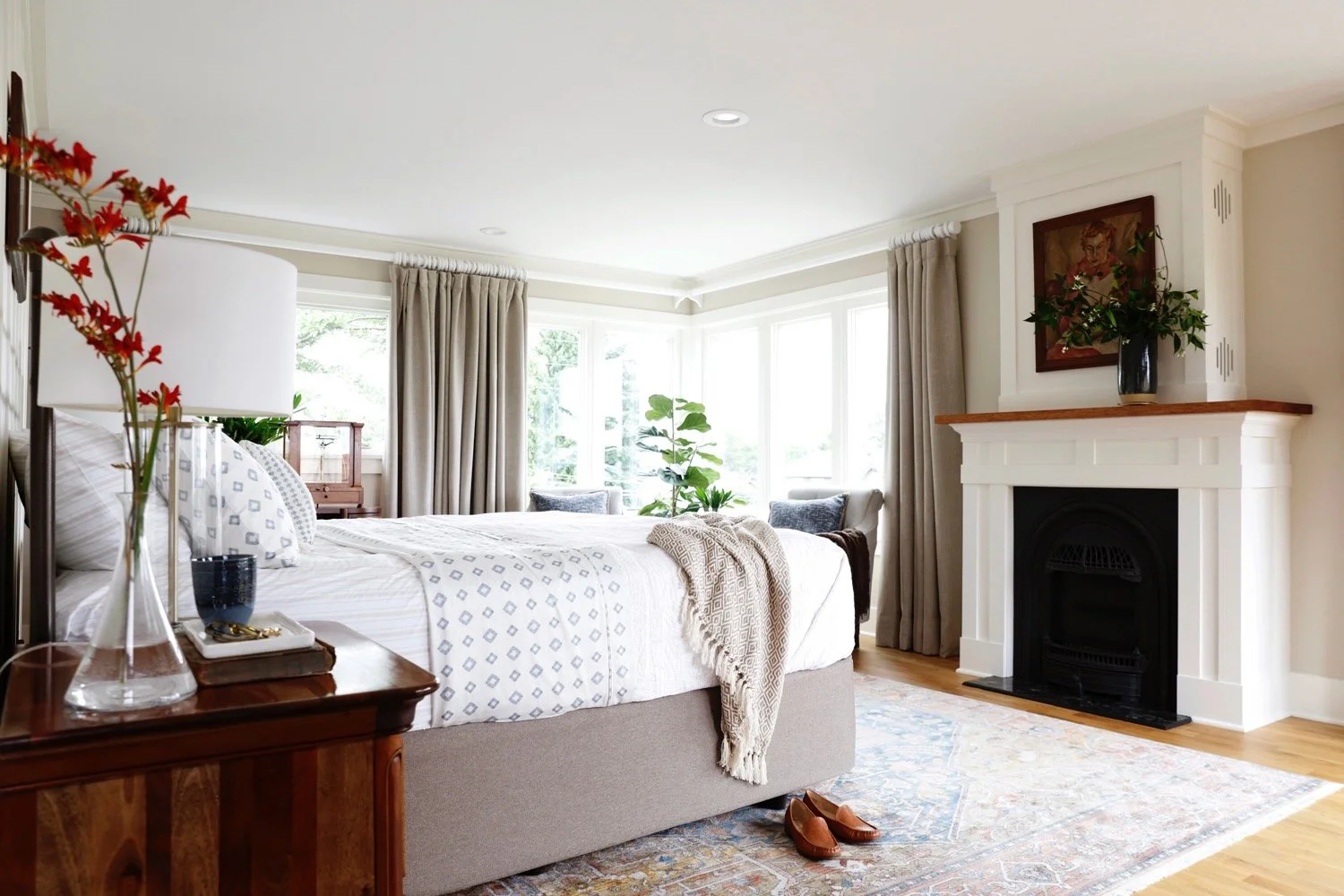 A bright bedroom with a large bed, white bedding, decorative pillows, a beige blanket, and brown slippers on a rug. There is a fireplace with a black insert, a vase with a plant on the mantel, an art piece above the fireplace, and windows with beige curtains. A small desk with a glass display case, a potted plant, and a nightstand with a lamp and glasses are also visible.