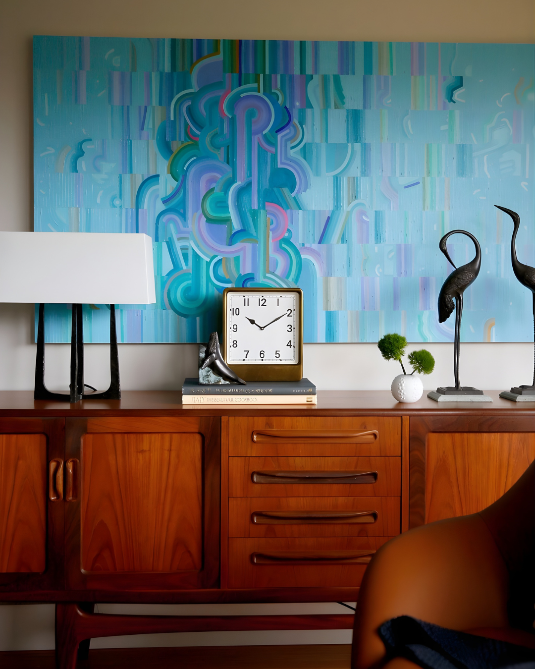 A mid-century modern wooden sideboard with a white wall clock, decorative black flamingo sculptures, a small white vase with green foliage, and a white lamp with a black base. Behind it is a large abstract blue and green painting.