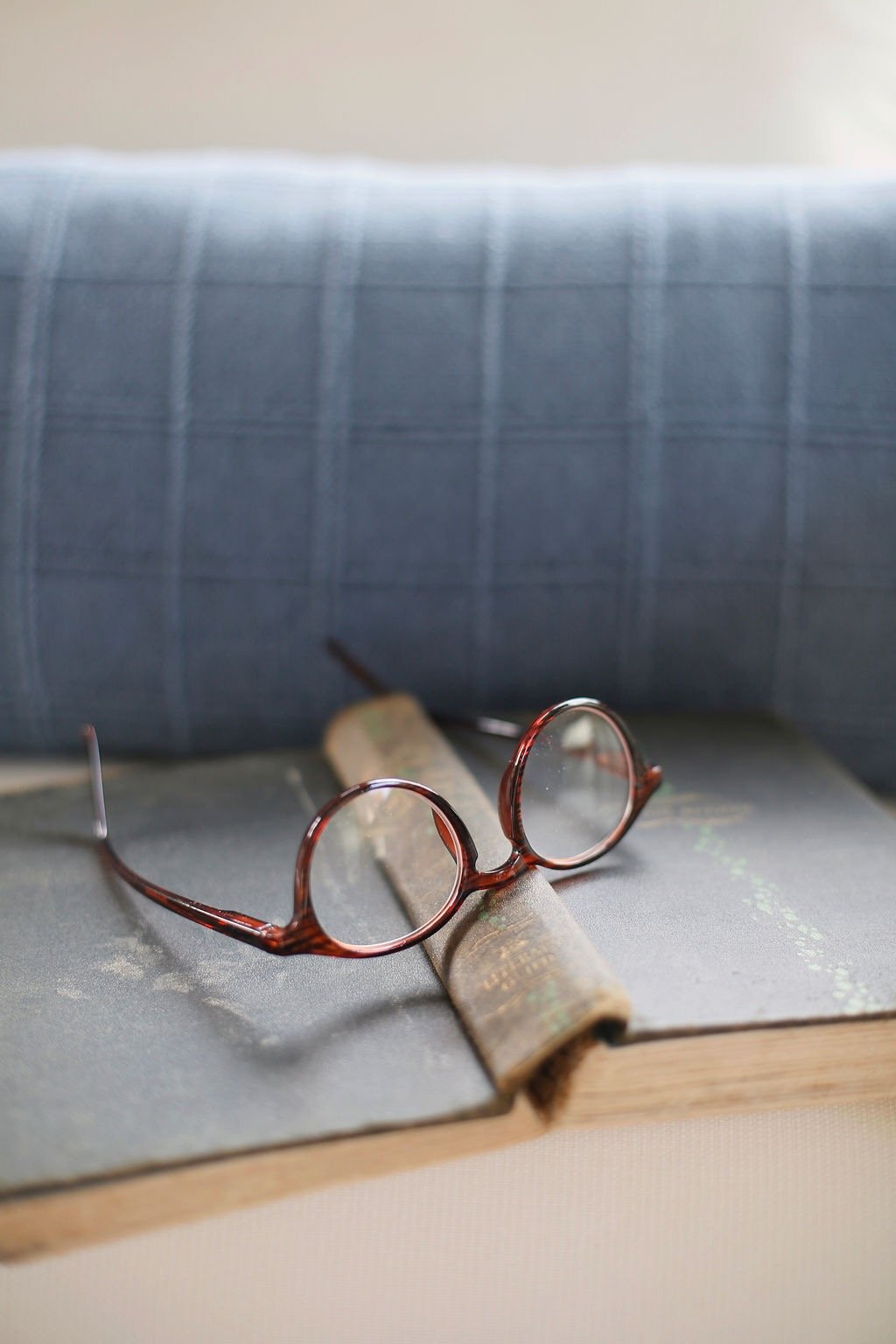 A pair of round eyeglasses with a tortoise shell frame placed on an open book, with a rolled-up newspaper underneath. In the background, there are two closed books stacked on top of each other.