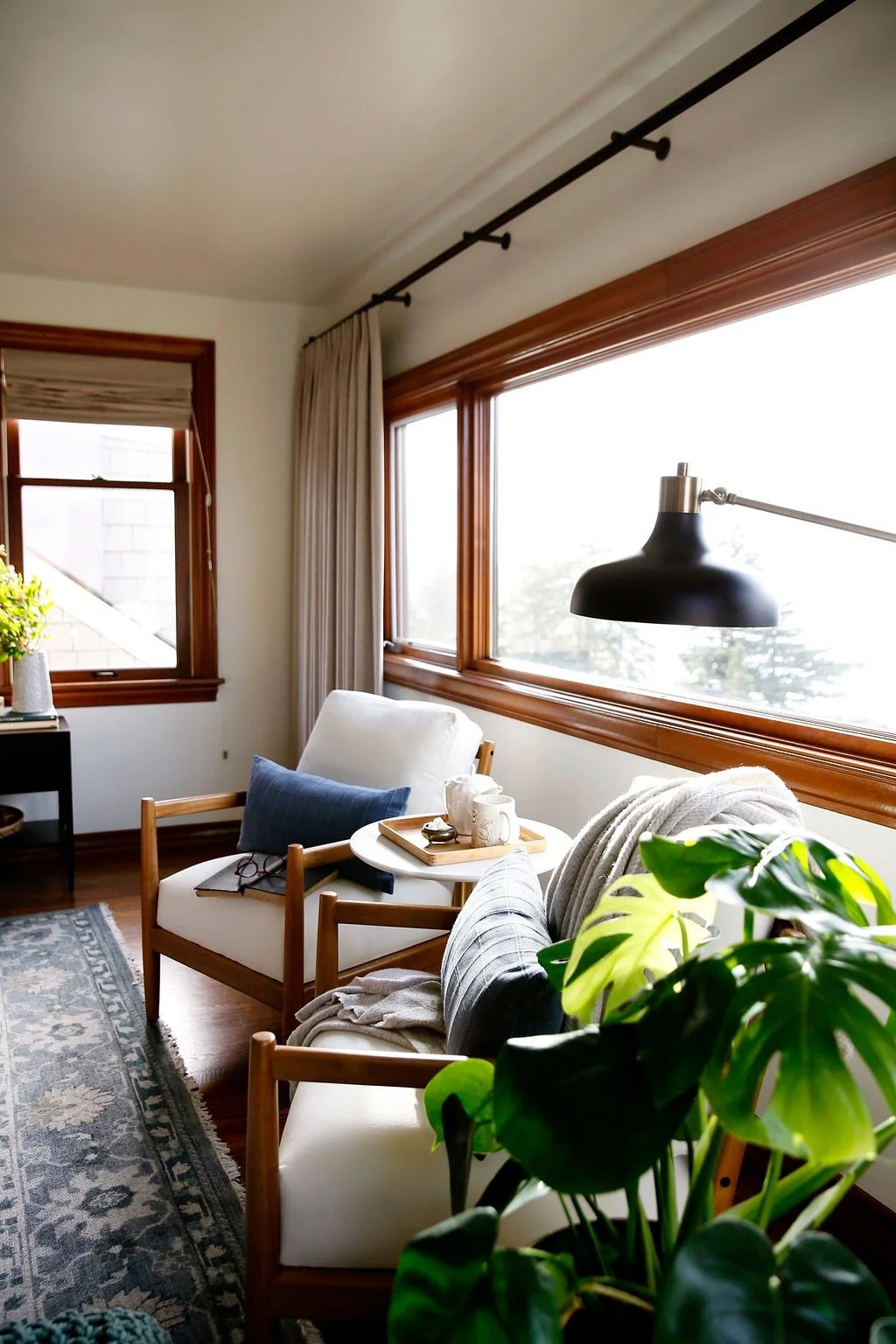 A cozy living room corner with large windows, a white armchair with a blue pillow, a side table with mugs and a tray, a black and white patterned rug, a dark wall lamp, and green indoor plants.