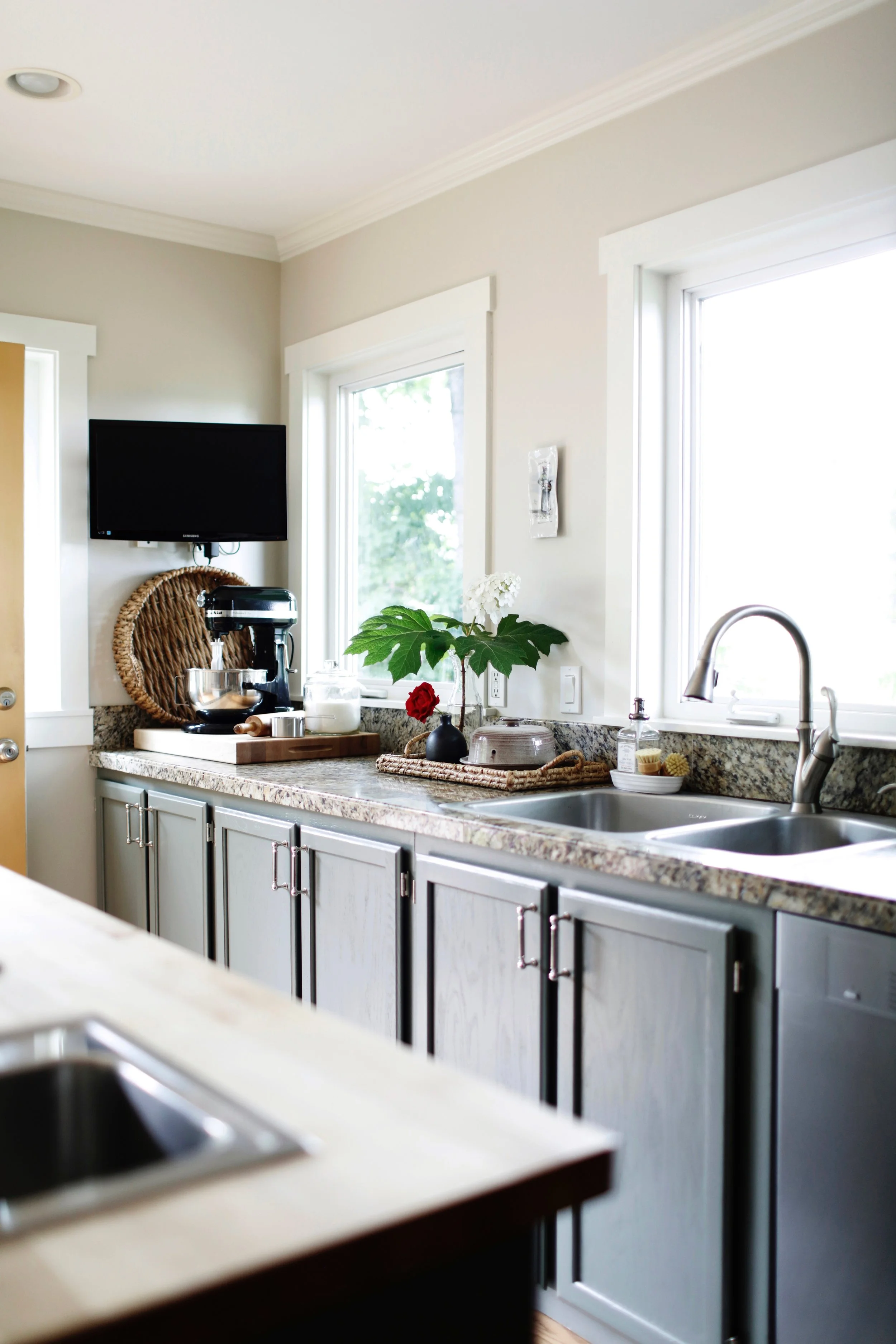 Bright kitchen with granite countertops, white cabinets, a stainless steel sink, plants, a small TV, and various kitchen items on the counter.
