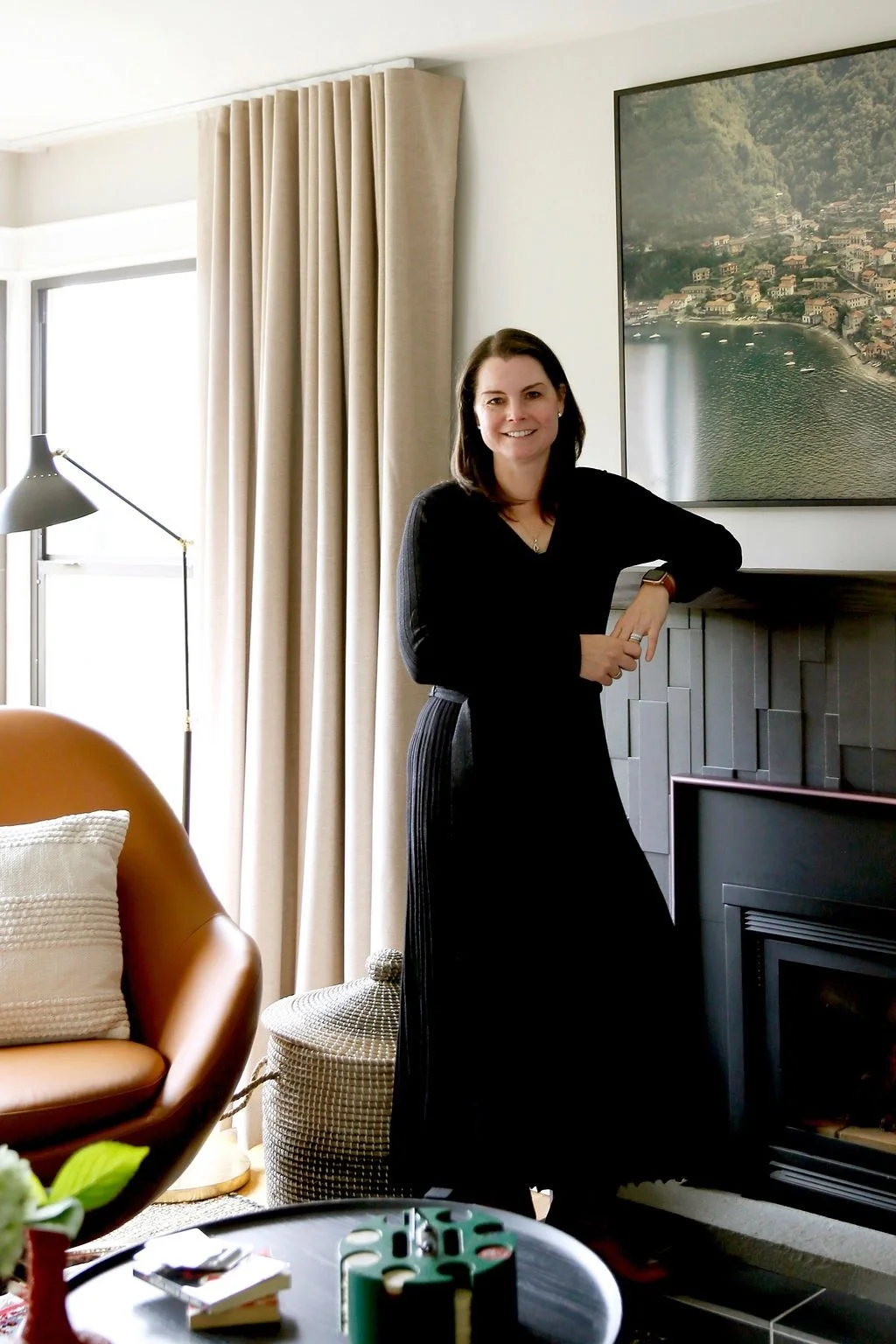 A woman with dark hair standing in a living room, leaning on a black fireplace mantel, smiling at the camera. Behind her is a large framed landscape photograph of a town by a lake, with beige curtains and a window on the left. In the foreground, there is an orange armchair with a white pillow, a round lamp, and a table with a green puzzle game and some books.