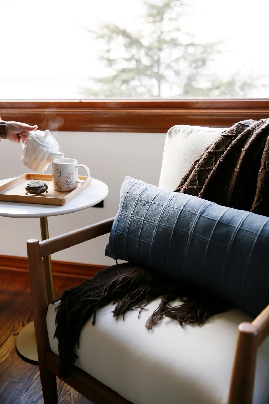 A cozy corner with a chair, a large pillow, and a throw blanket, with hot tea being poured into a mug on a side table by a window overlooking trees.