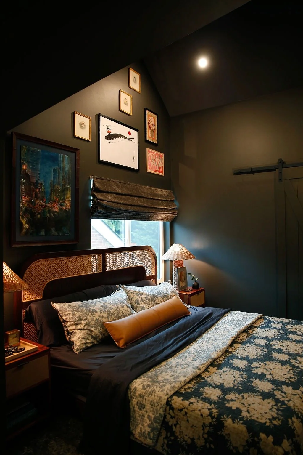 A cozy bedroom with a bed that has a black and beige floral bedspread, multiple pillows, wooden nightstands with lamps, and artwork on the dark-painted walls, including a window with a brown Roman shade and a full moon visible outside.