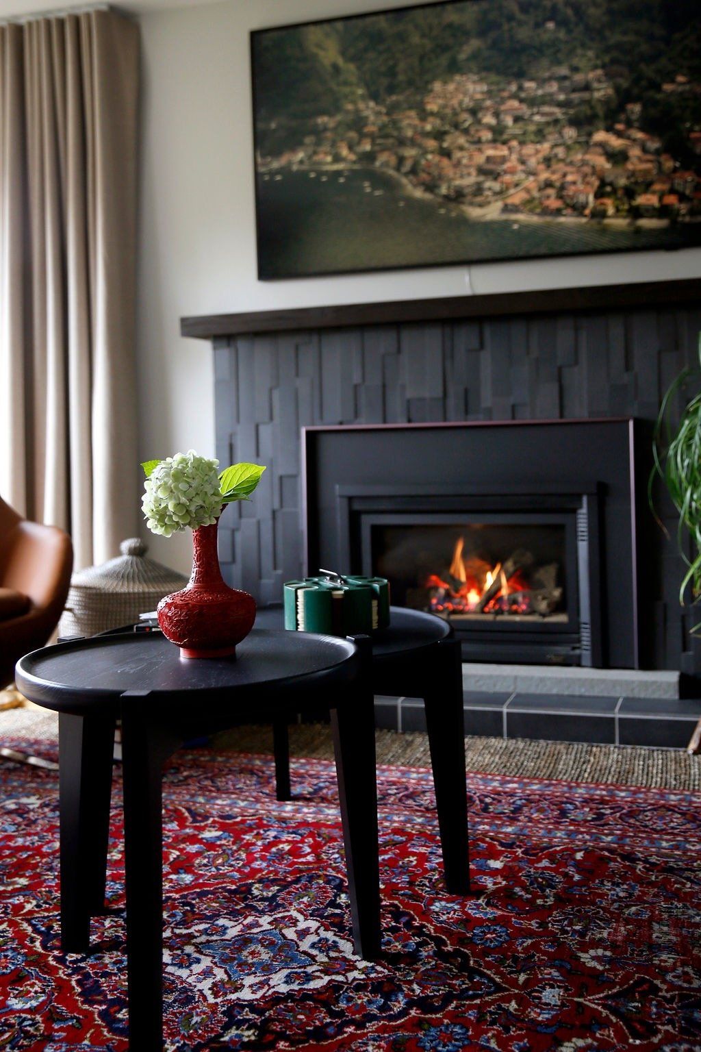 Living room with a fireplace, a black coffee table with a red vase and green candles, a patterned rug, and a large landscape painting above the fireplace.