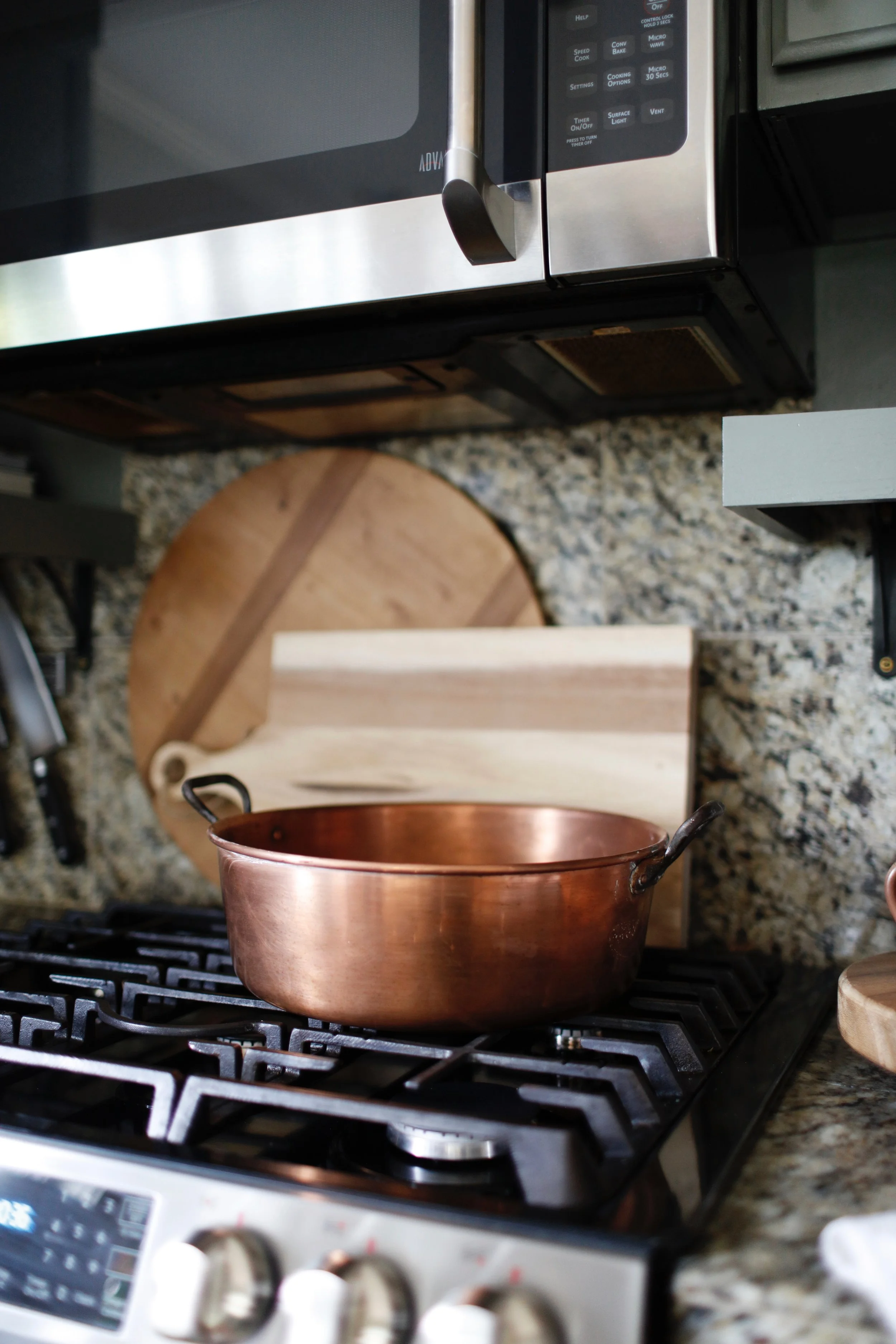 A copper pot on a stove with a granite countertop, a wooden cutting board, and a microwave above it.