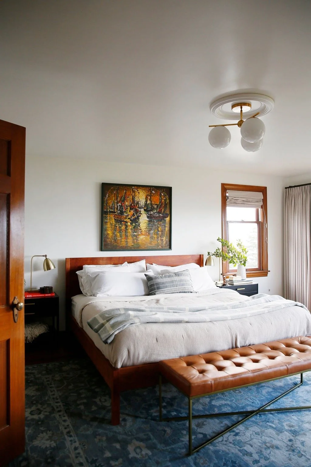 A cozy bedroom featuring a wooden bed frame with white bedding and a gray plaid pillow. There's a bench at the foot of the bed, a nightstand with a lamp, a window with wooden trim, a piece of artwork above the bed, and a ceiling light with three globe bulbs.
