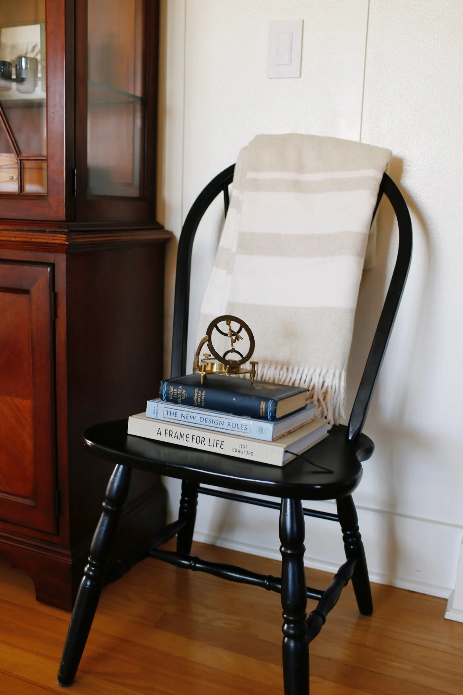 A black wooden chair with a beige and white striped towel draped over the backrest. On the seat of the chair are four stacked hardcover books, with titles visible: "The New Design Rules," "A Frame for Life," and others. To the left of the chair, ther