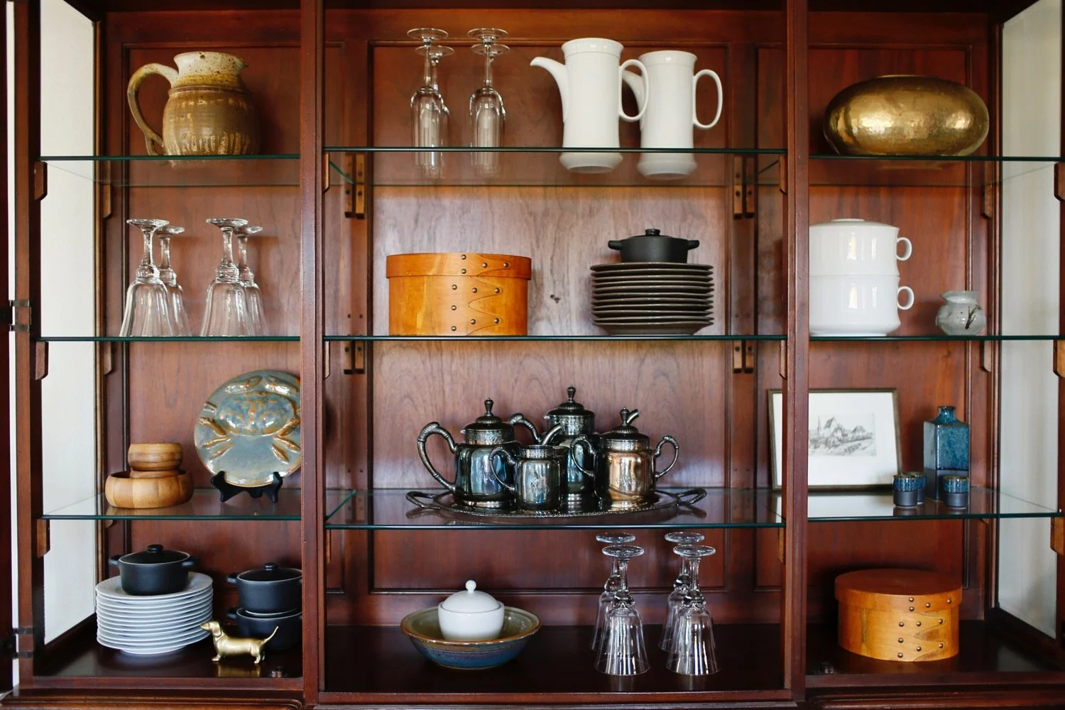Wooden display cabinet with glass shelves holding dishes, glassware, and decorative items.