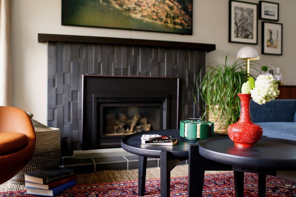 Living room with fireplace, two black coffee tables, red vase with white flowers, green plant, books, and framed artwork on the wall.