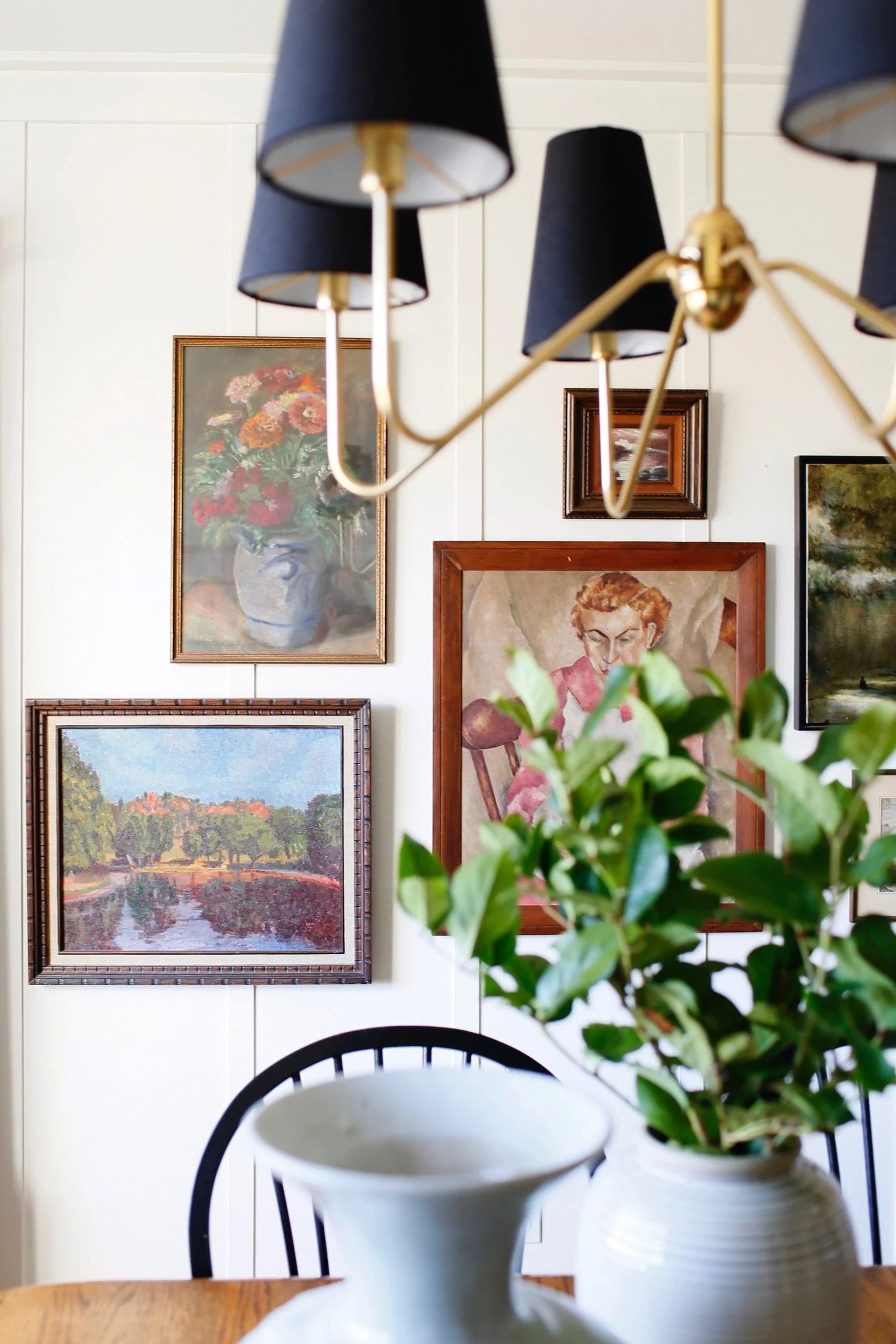 Interior of a room with a wall decorated with framed paintings, a wooden table, and a black dining chair. In the foreground, there are two white vases on the table, one containing a green leafy plant. A black chandelier with black lampshades is hangi