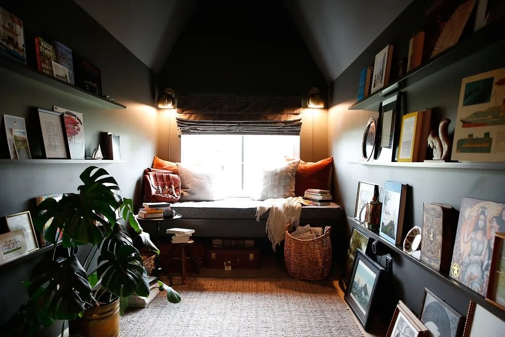 Cozy attic room with a window seat, surrounded by shelves of books and artwork, with a large houseplant in the foreground.